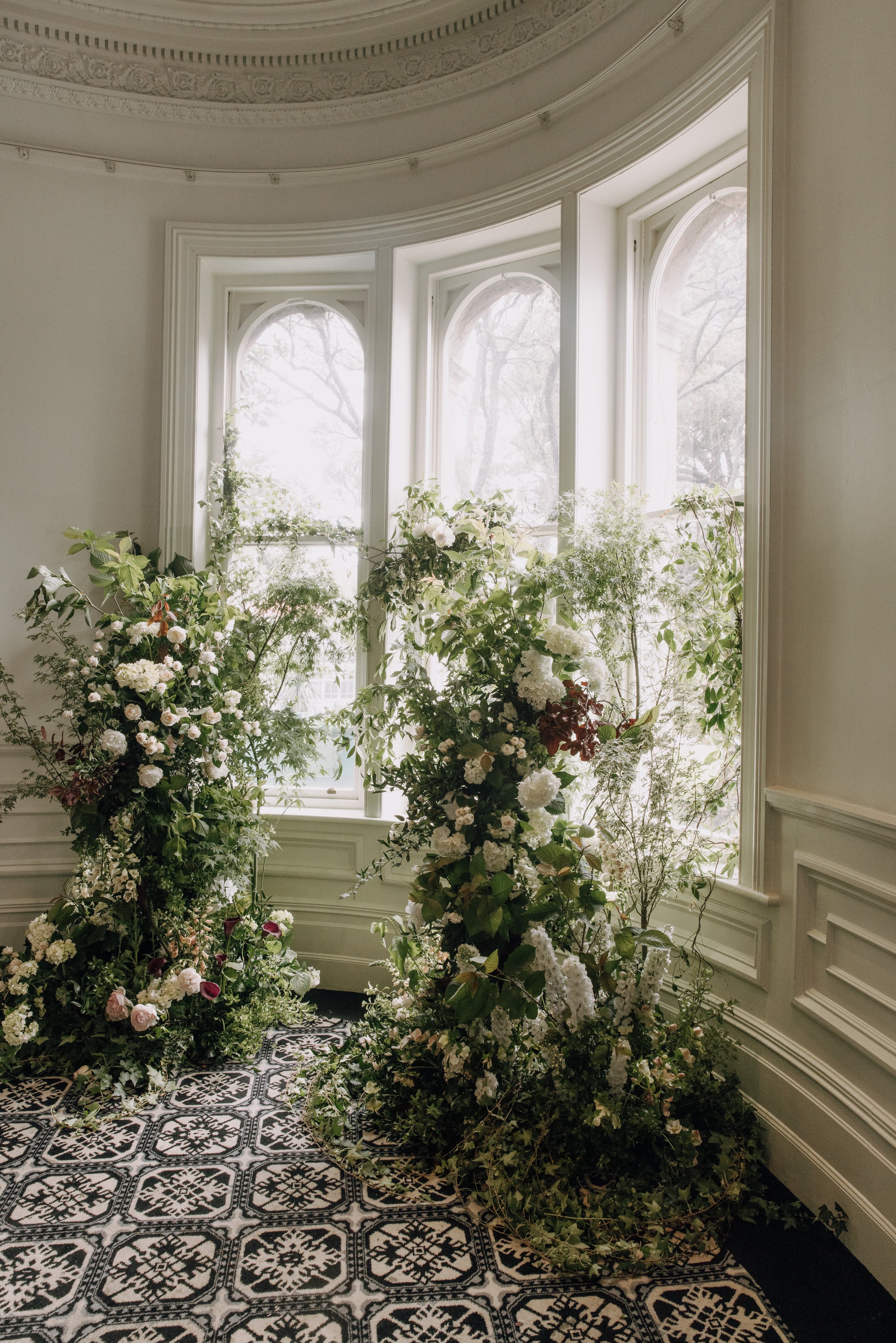 A bright room with large bay windows filled with sunlight and surrounded by lush green plants and flowers, on a patterned black and white tile floor, with decorative molding on the walls.