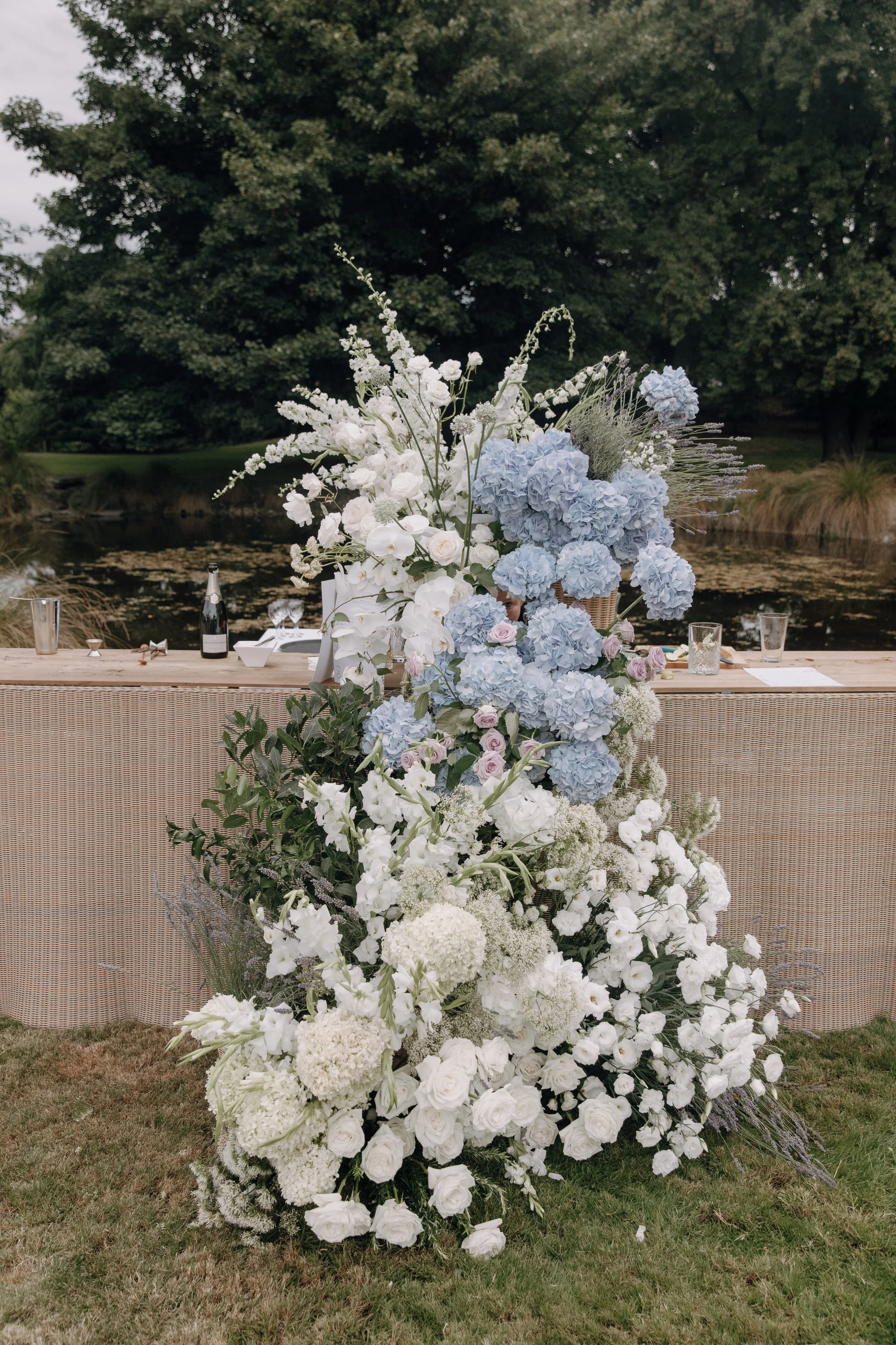 A large floral arrangement with white and light blue flowers displayed outdoors near a body of water, with a table set behind it, on a grassy area during daytime.