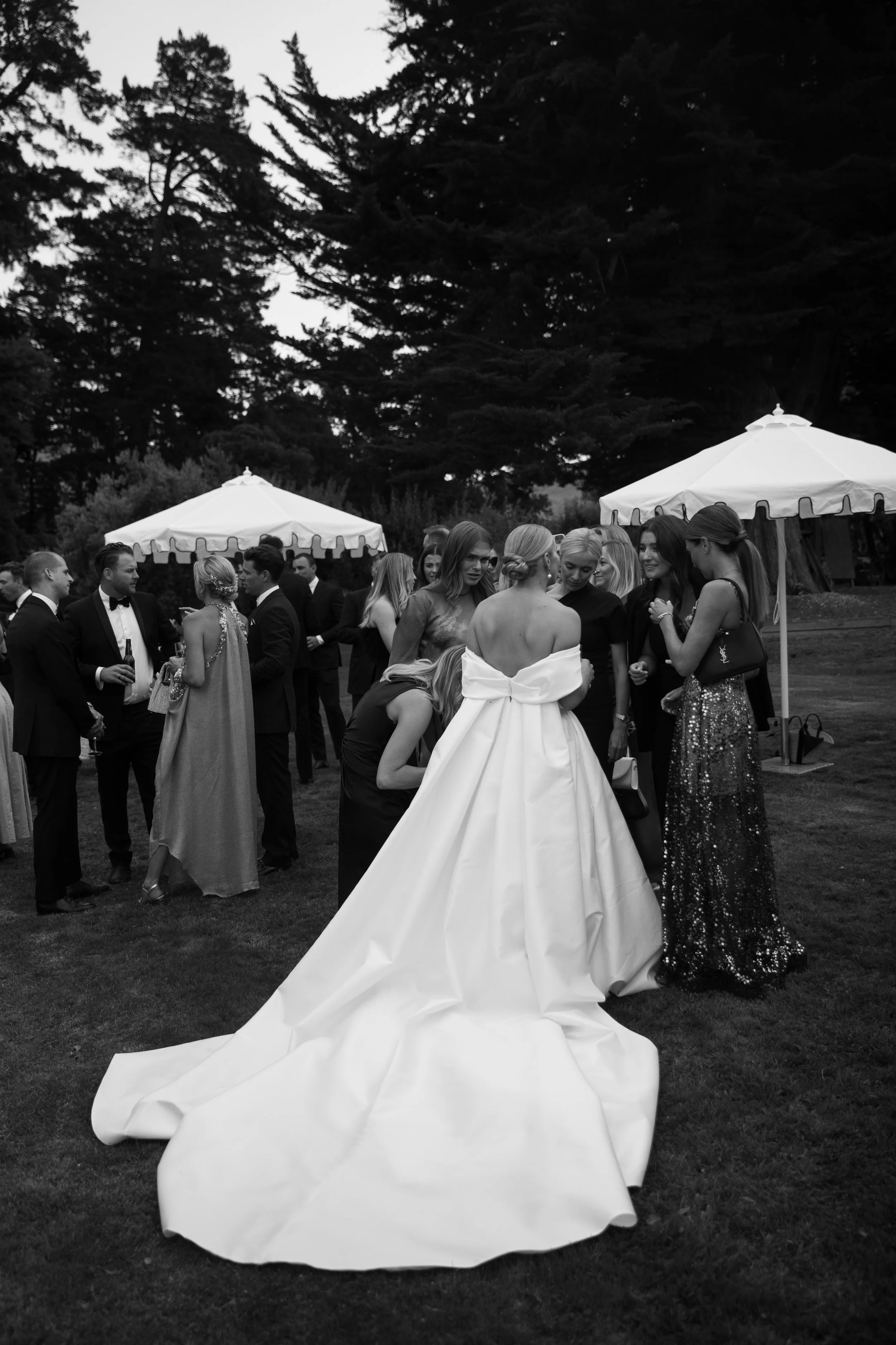 Black and white photograph at an outdoor event with guests dressed in formal attire, including a woman in a wedding gown and a group of women chatting near a tent, with tall trees in the background.