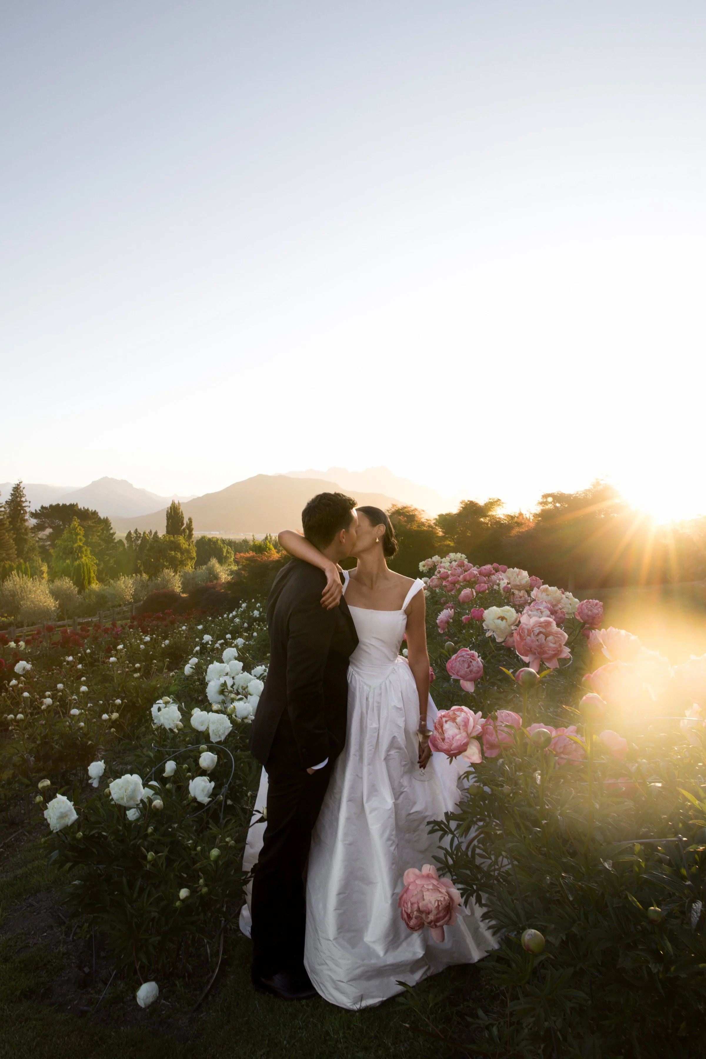 A couple dressed in wedding attire sharing a kiss in a garden filled with pink and white flowers during sunset.