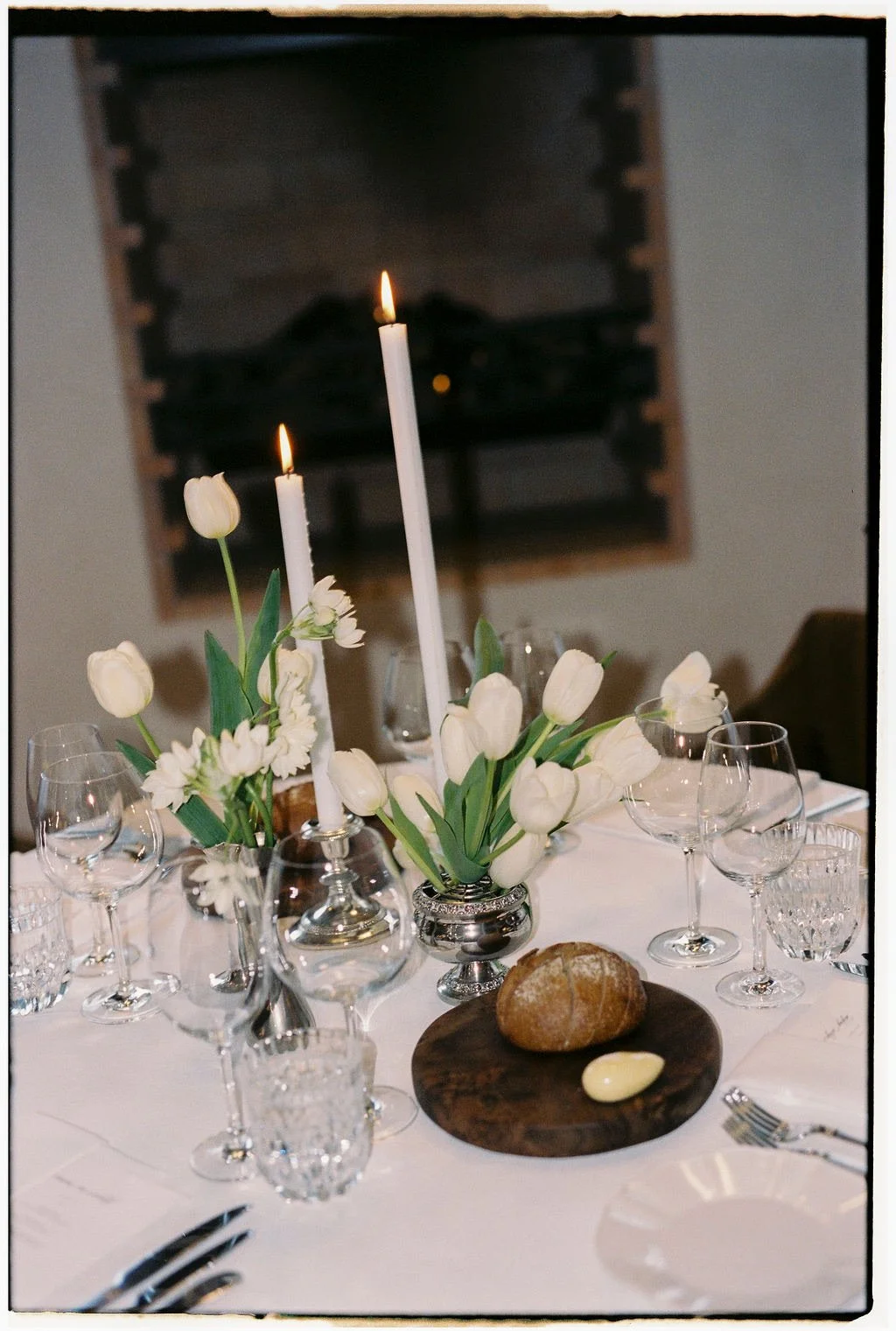 Elegant table setting with white tulips and candlesticks, bread on a wooden board, and multiple wine glasses.