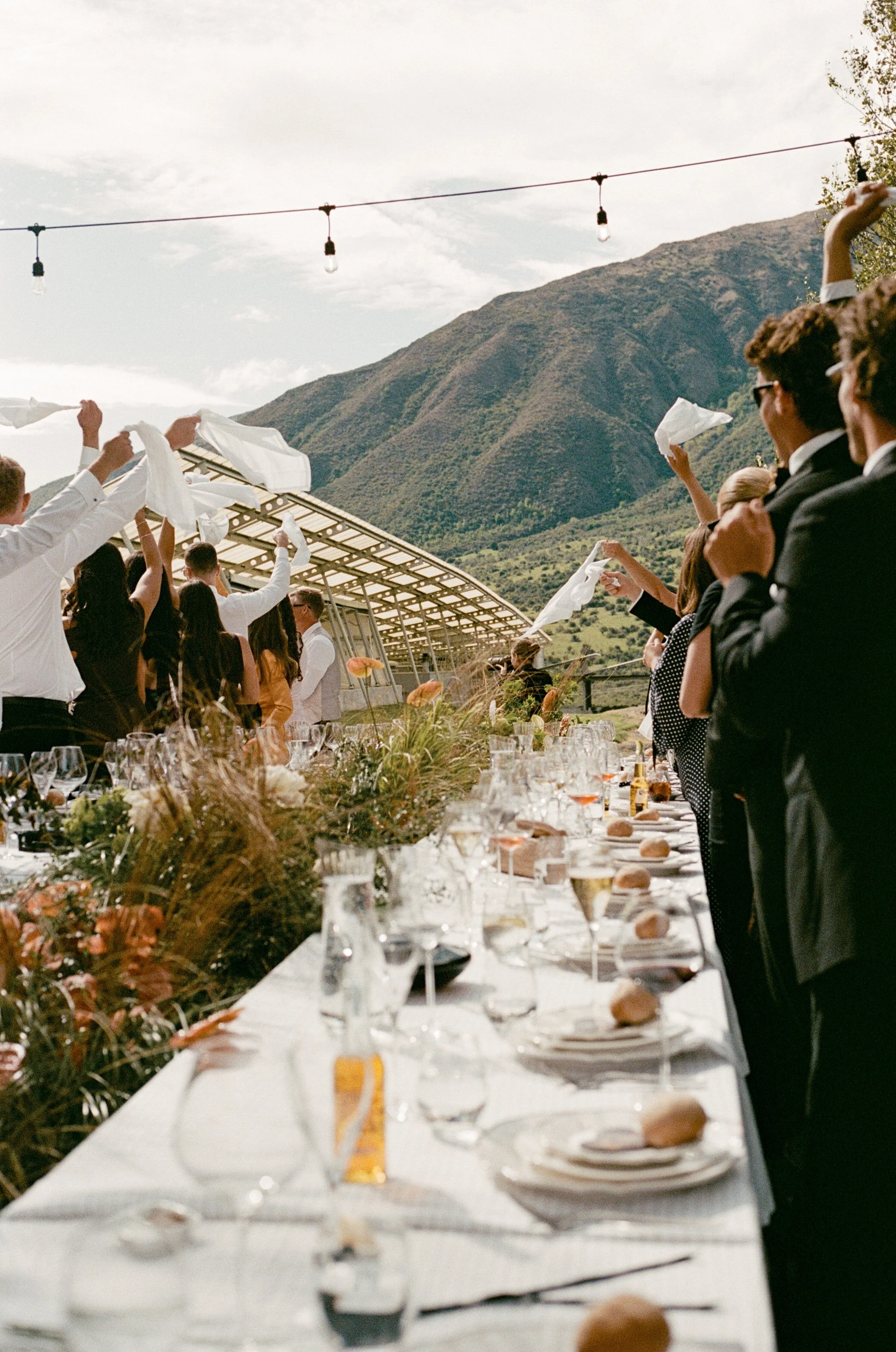 People celebrating at an outdoor event with mountains in the background, tables set with white tablecloths, glassware, and food, and individuals waving napkins in the air.
