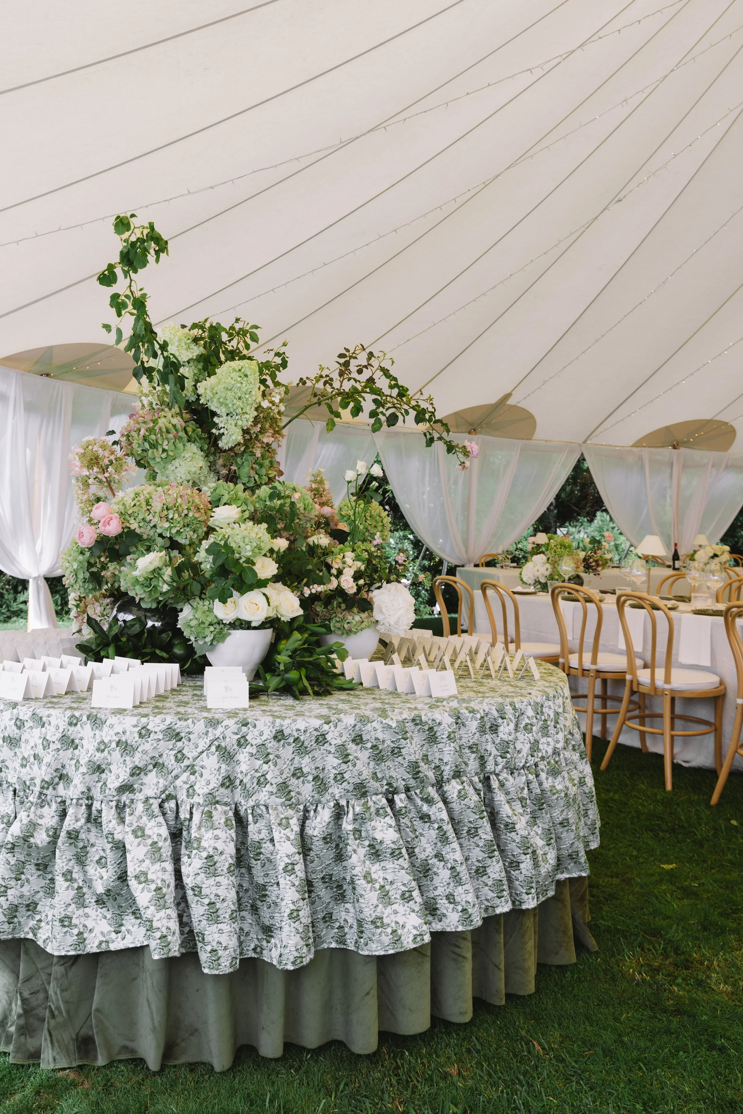A large floral centerpiece with pink, green, and white flowers on a table decorated with a green floral tablecloth at an outdoor event under a white tent.