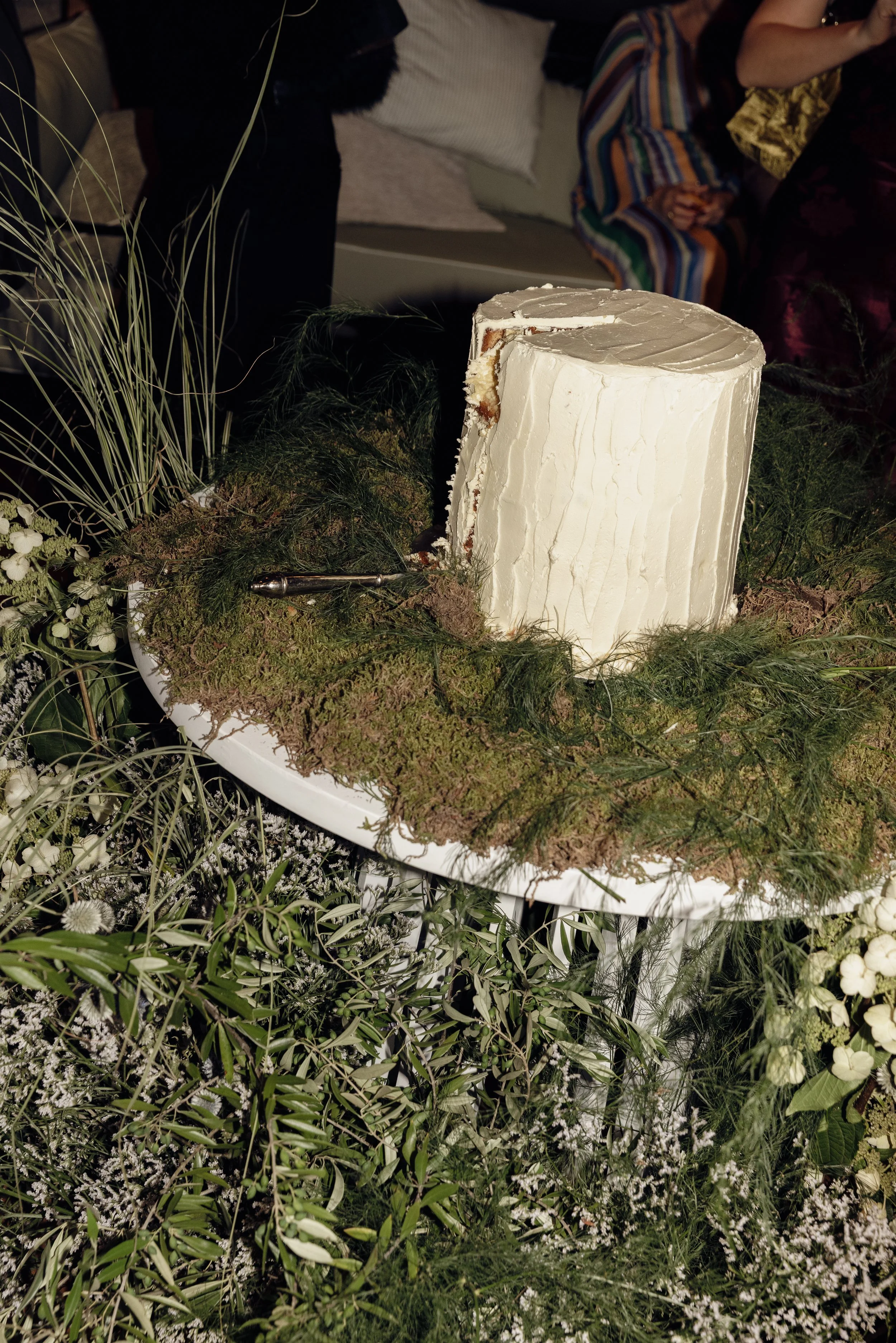 A large white cake with smooth icing and decorative swirls on top, placed on a table decorated with greenery and white flowers for a celebration.