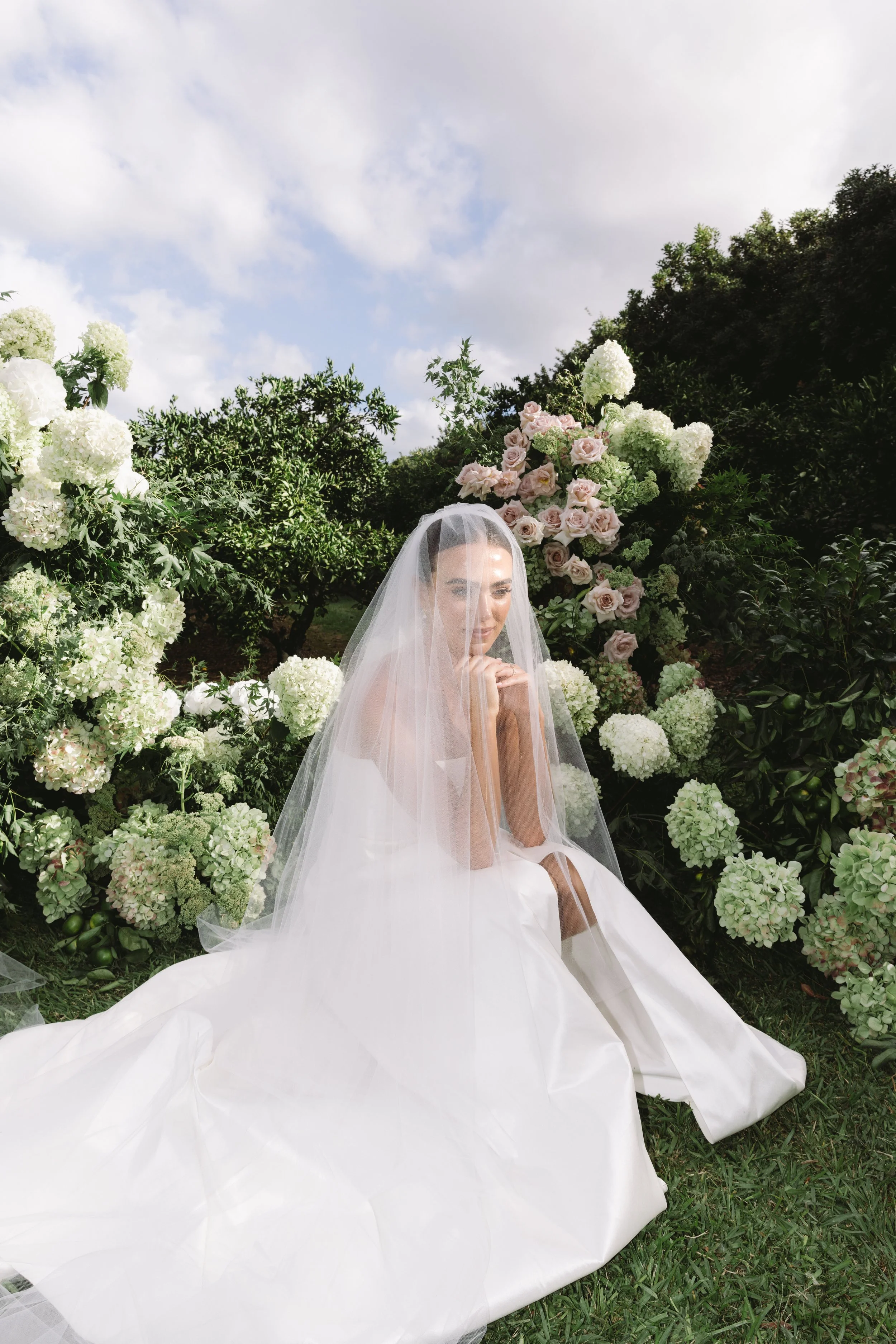 A bride sitting outdoors among white and light pink roses and hydrangeas, dressed in a wedding gown with a veil, under a partly cloudy sky.