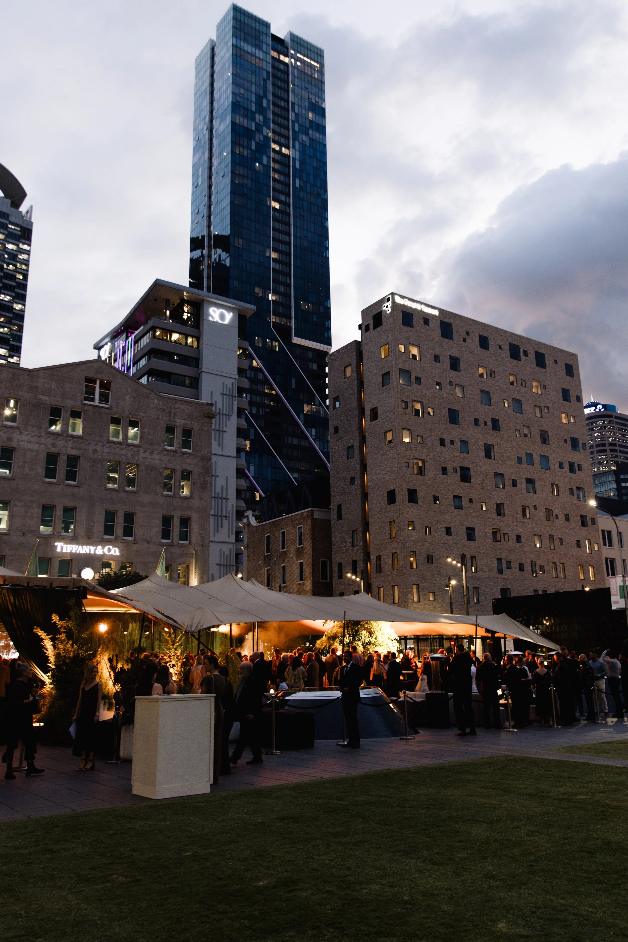People attending an outdoor event with tents and string lights in a cityscape at dusk, with tall modern buildings and cloudy sky in the background.