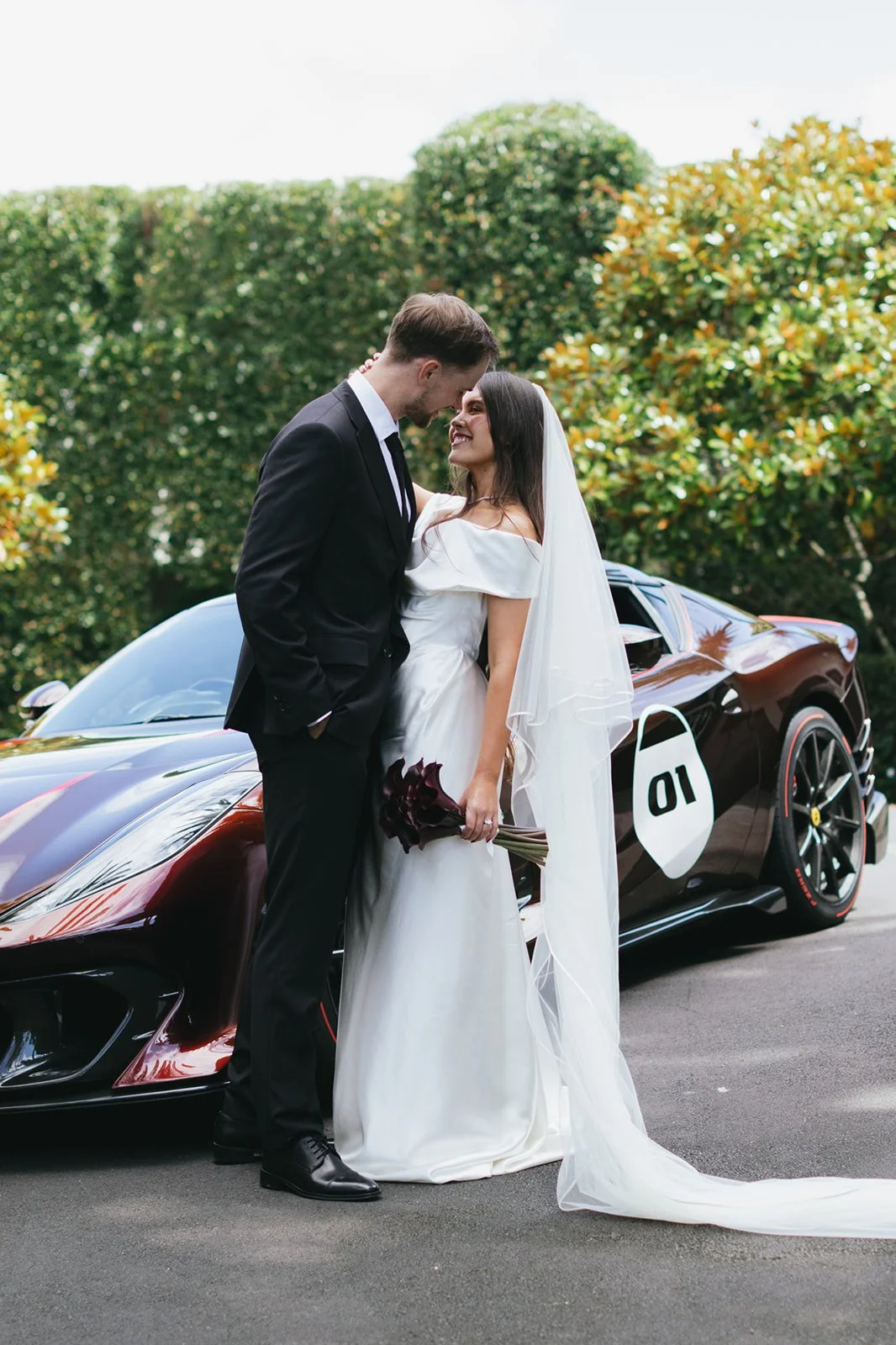 A bride and groom standing close together in front of a race car, smiling at each other. The bride holds a bouquet of dark flowers, and the groom is dressed in a black suit. The scene is outdoors with greenery in the background.