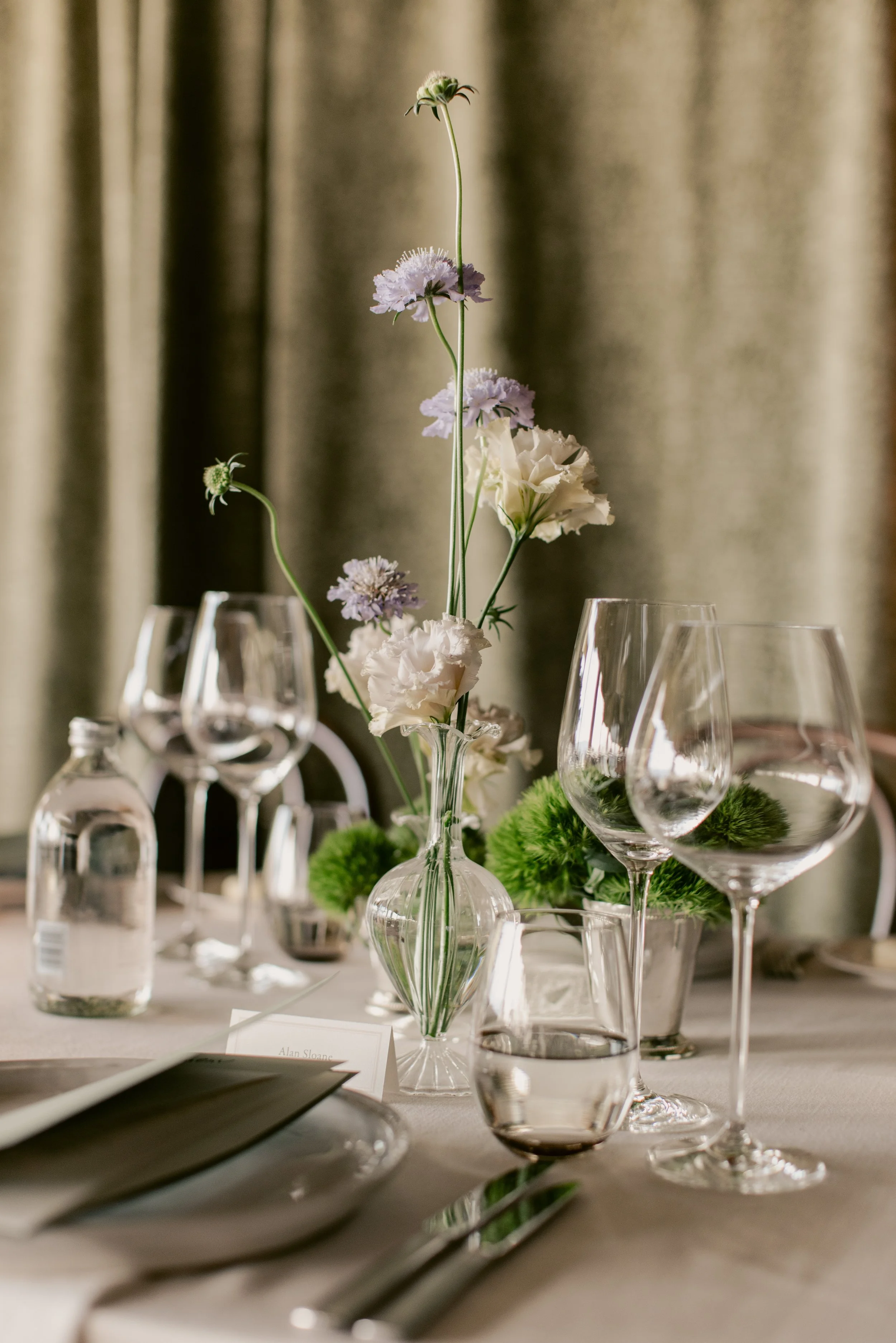 A table setting with a minimalist floral centerpiece, clear glassware, and a blurred background of beige curtains.