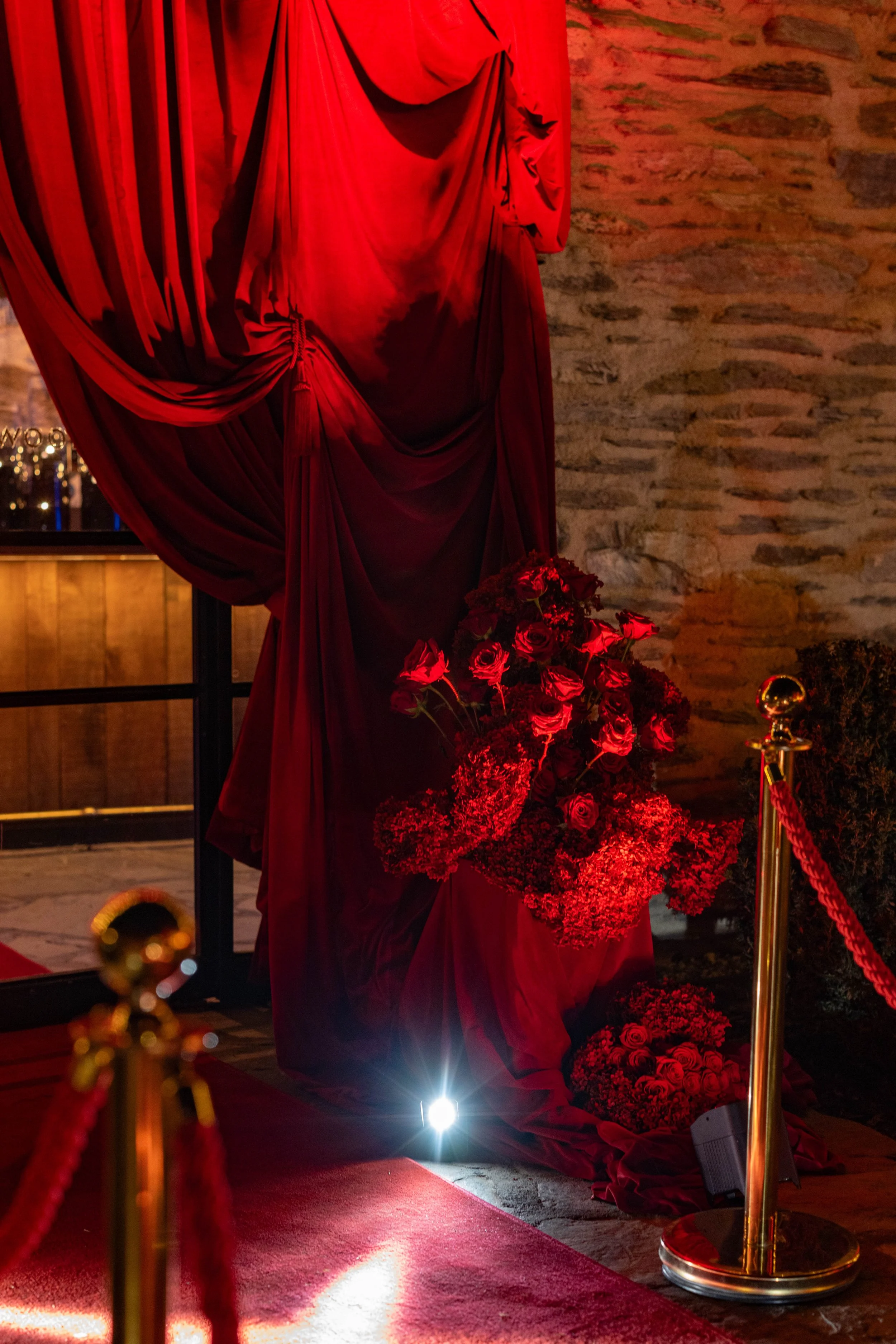 A display of red roses and a red draped curtain, roped off with gold stanchions and red velvet ropes, illuminated by spotlight, in front of a brick wall.