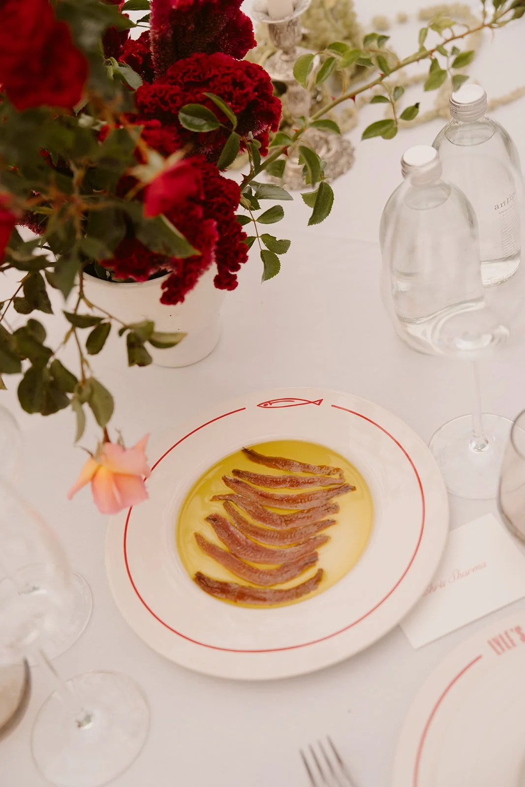 A table with a white plate containing small strips of bacon in yellow oil, surrounded by glasses, water bottles, and a bouquet of red and pink flowers.