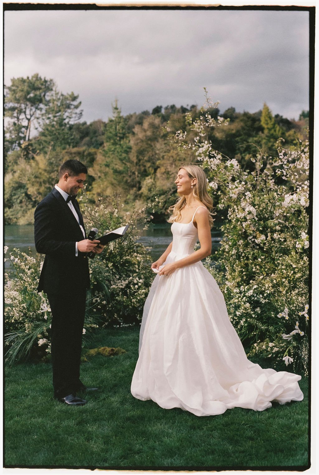 A bride and groom exchanging vows outdoors surrounded by blooming white flowers and greenery, with a river and trees in the background.
