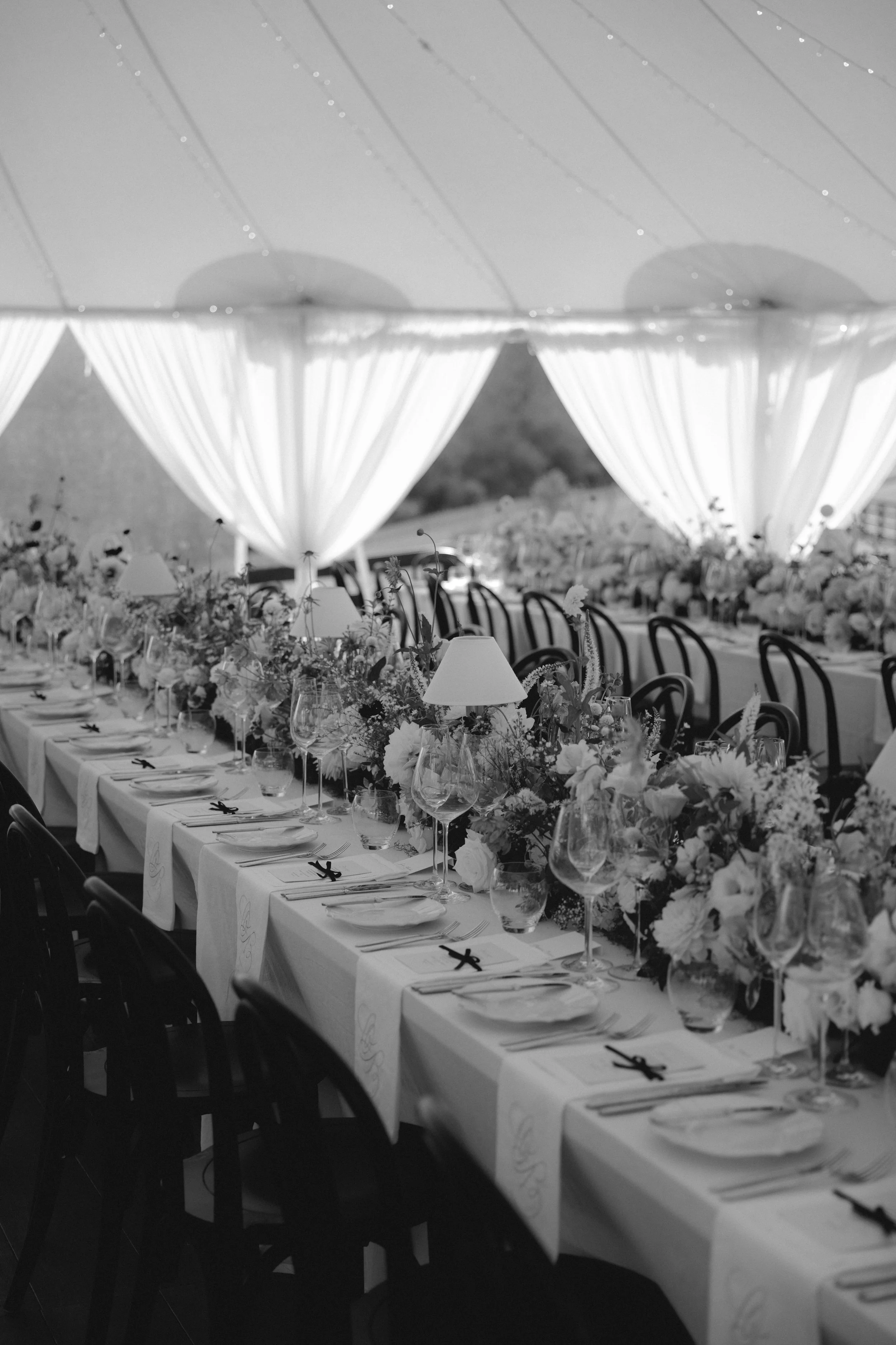 A long banquet table decorated with flowers, wine glasses, and place settings under large, draped white curtains at an outdoor event.