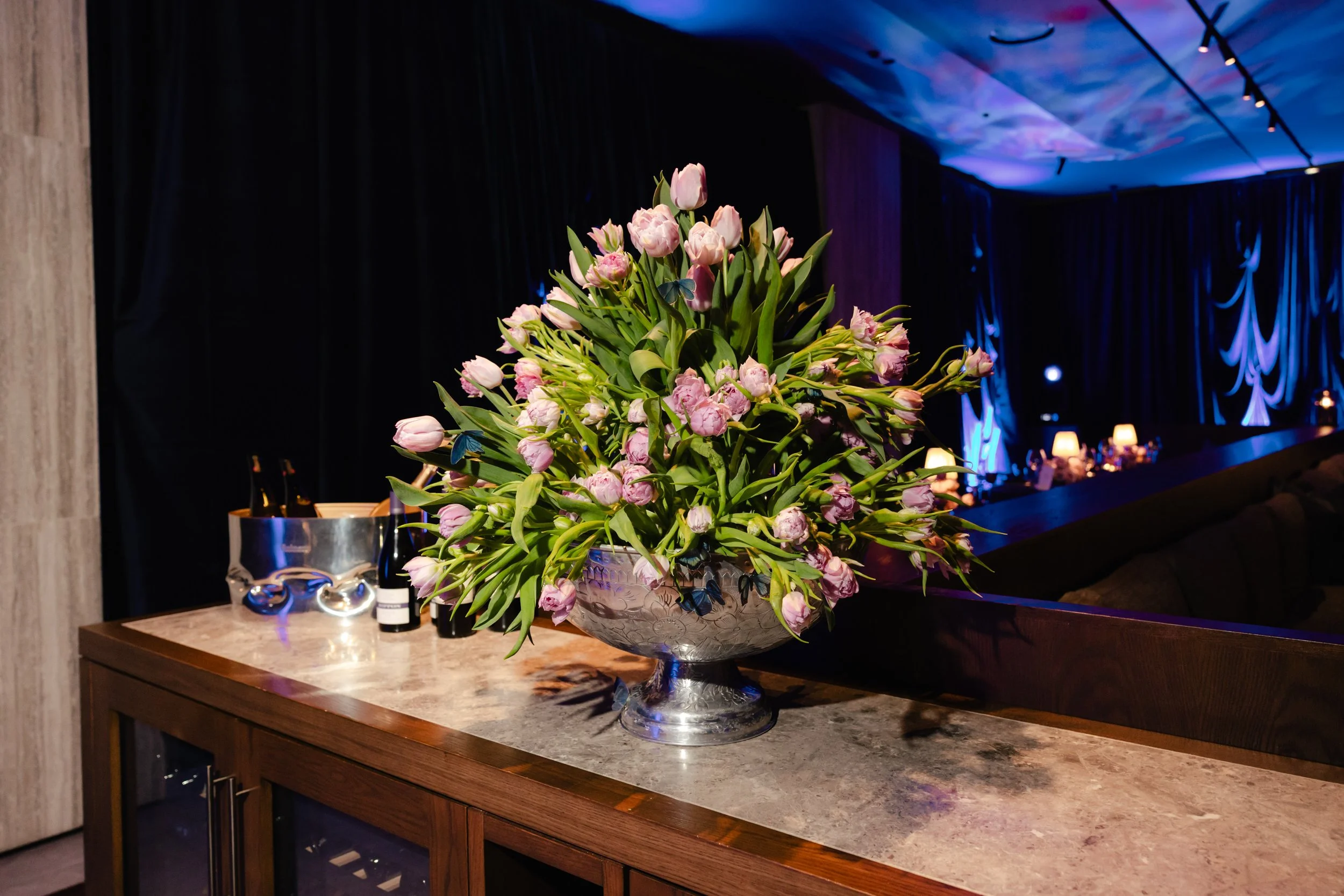 A large bouquet of pink tulips with green leaves is arranged in a silver decorative bowl on a wooden counter. In the background, there are dim blue and purple lighting candles and tables, suggesting a festive or elegant event setting.