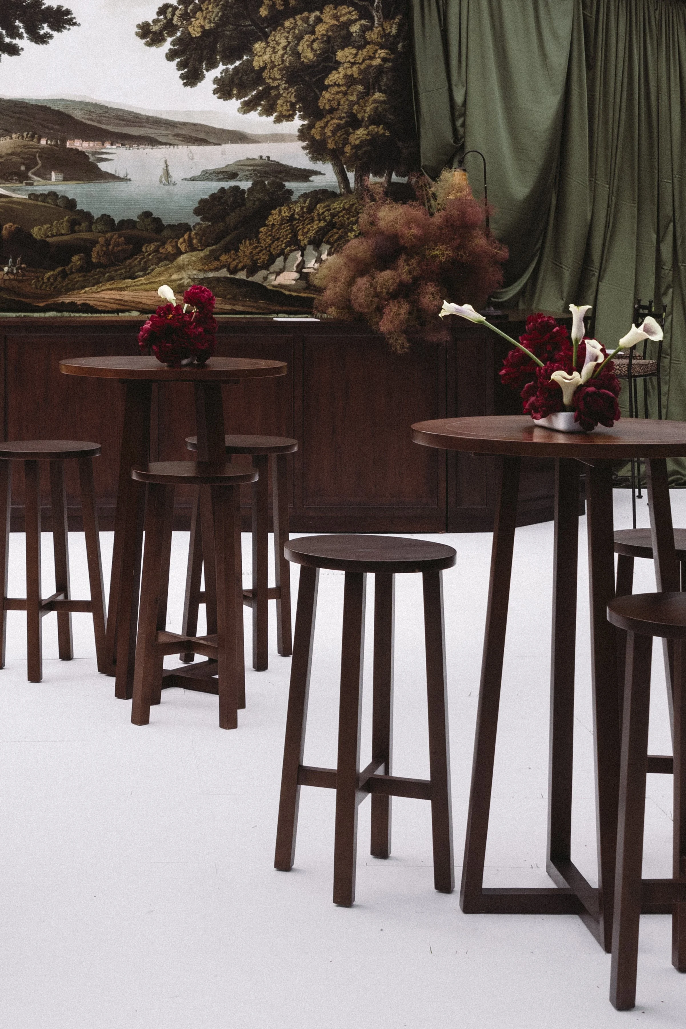 Interior scene with wooden tables and stools, floral arrangements, and a large mural of a landscape with water, trees, and islands on the wall.