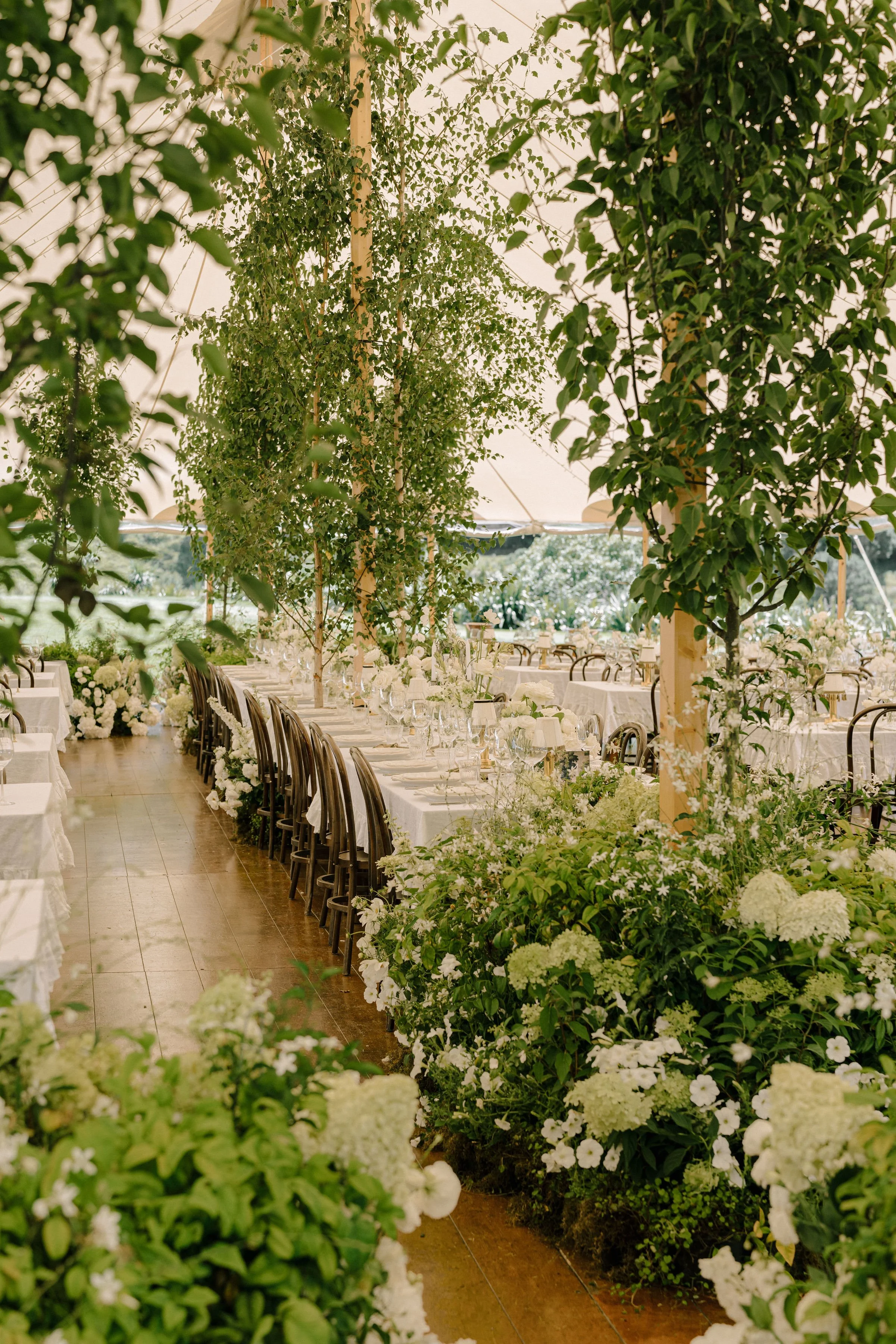 Long banquet table decorated with white flowers and greenery, under a tent ceiling with large green trees surrounding it.