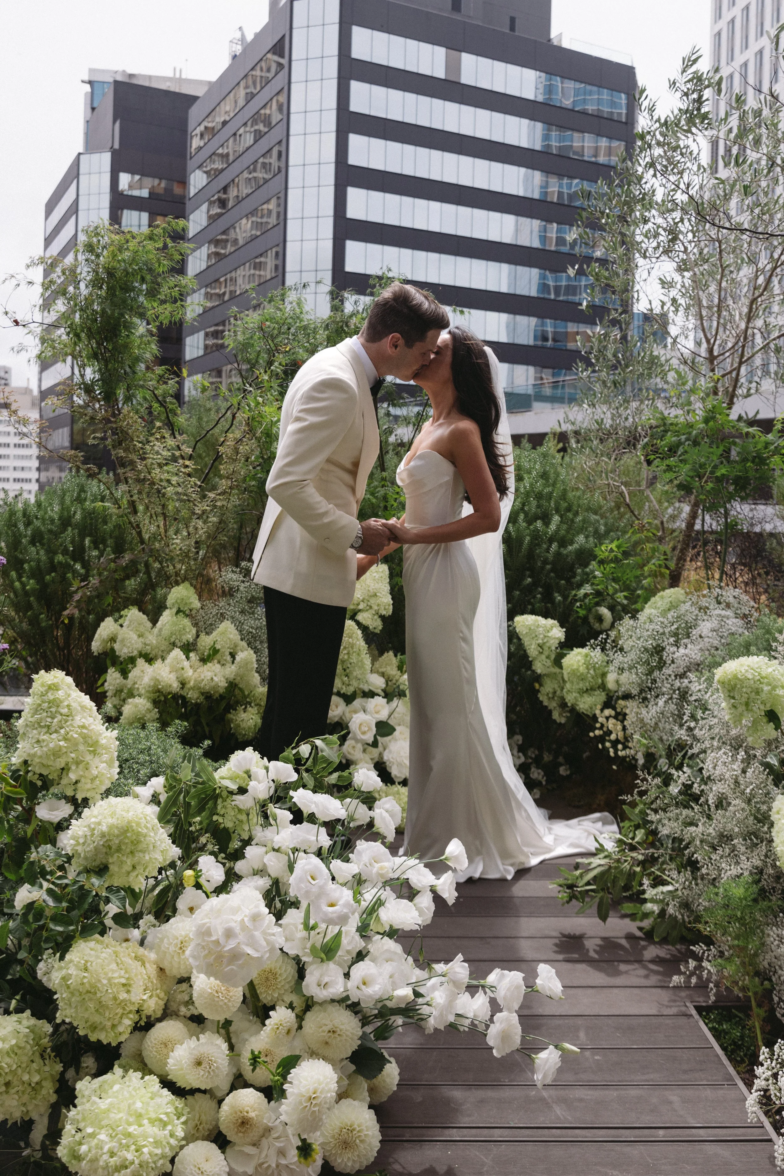 A couple dressed in wedding attire sharing a kiss on a floral outdoor platform in an urban cityscape with modern buildings and trees.