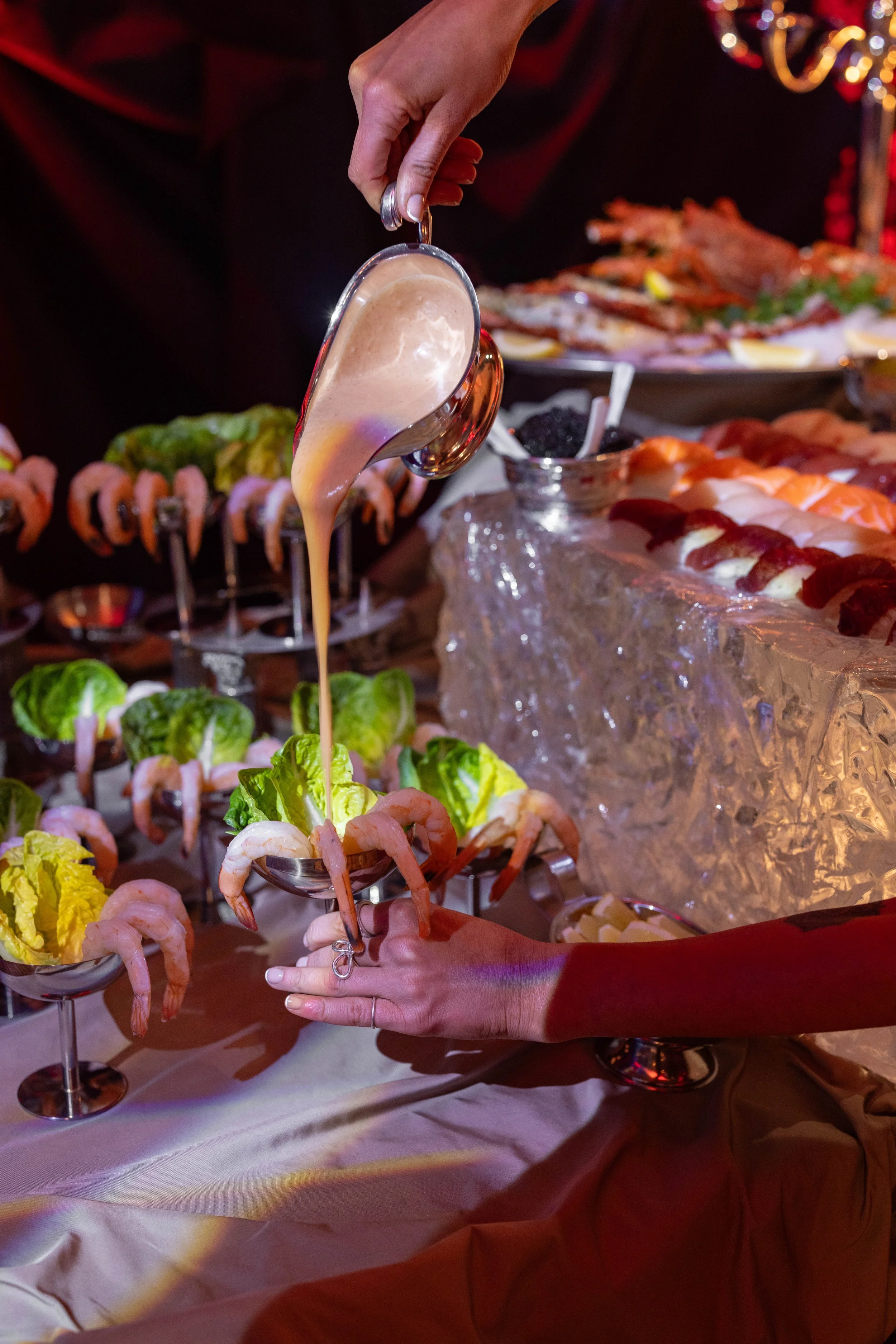 A hand holding a small metal cup filled with shrimp, with sauce being poured into it at a seafood buffet, surrounded by other seafood dishes and garnishes.