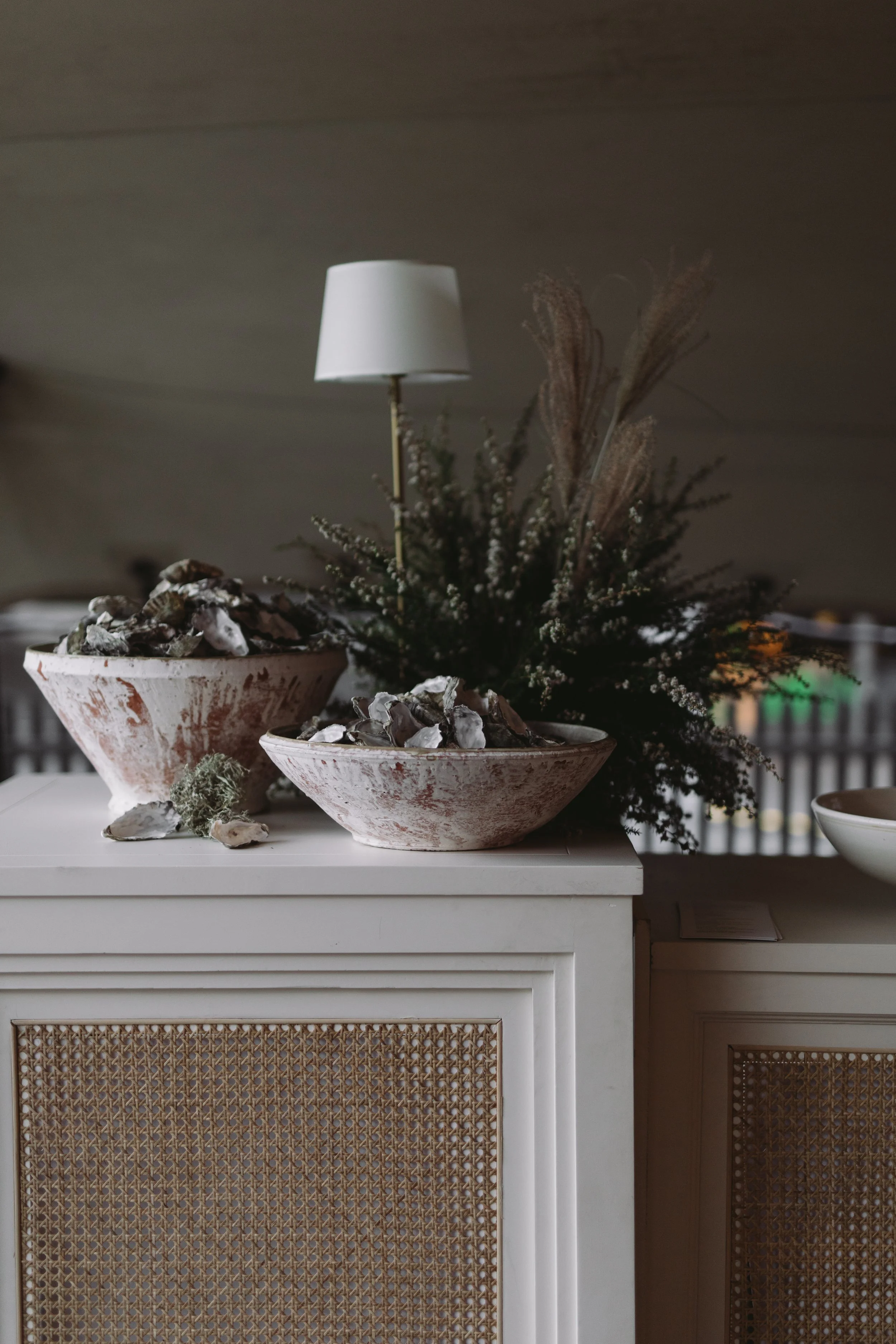 Decorative display with large white bowls filled with seashells. A white table with a rattan-panel door design is in front, topped with a floral arrangement and a small lamp.