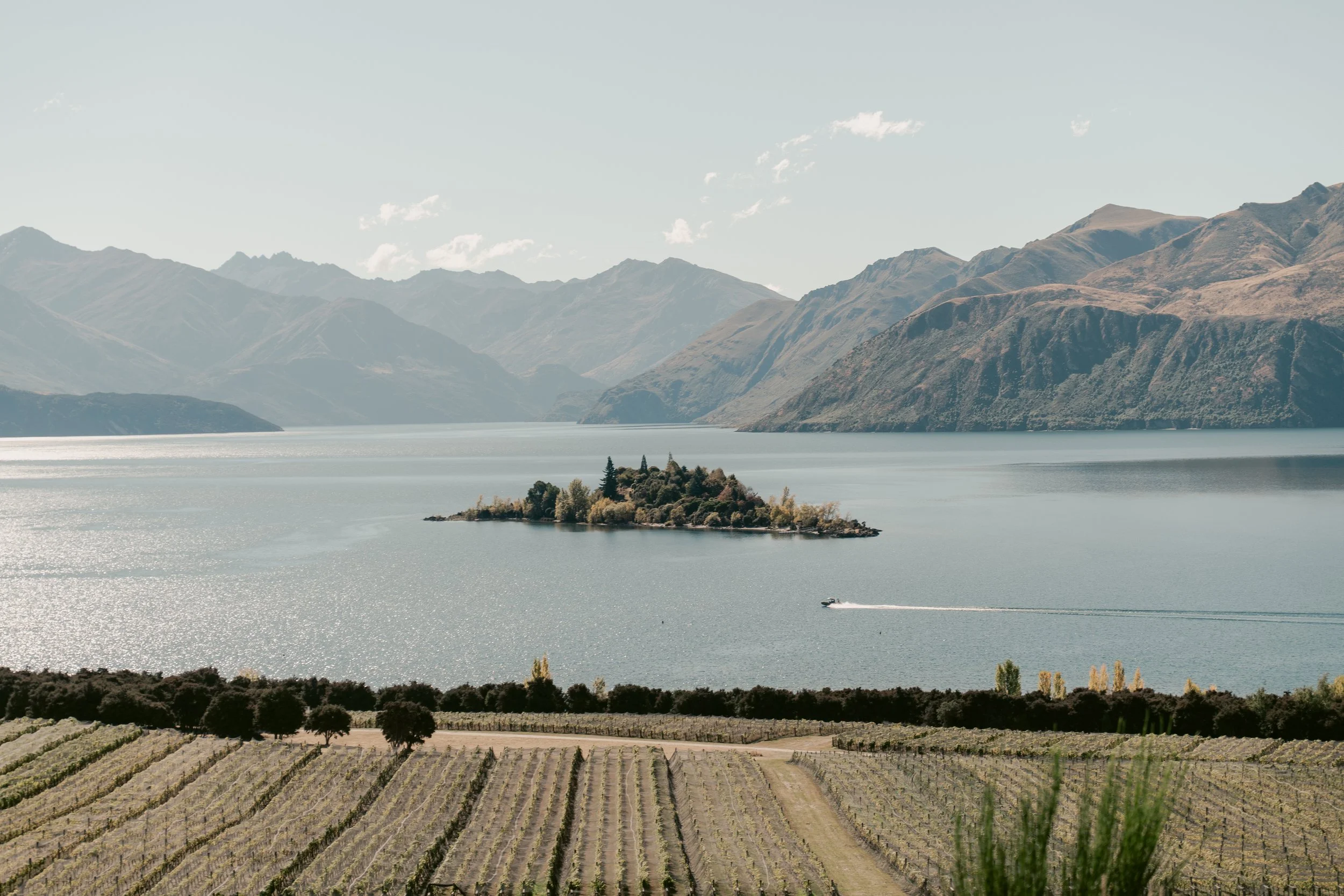 A large body of water with a small forested island in the middle, surrounded by mountain ranges under a partly cloudy sky.