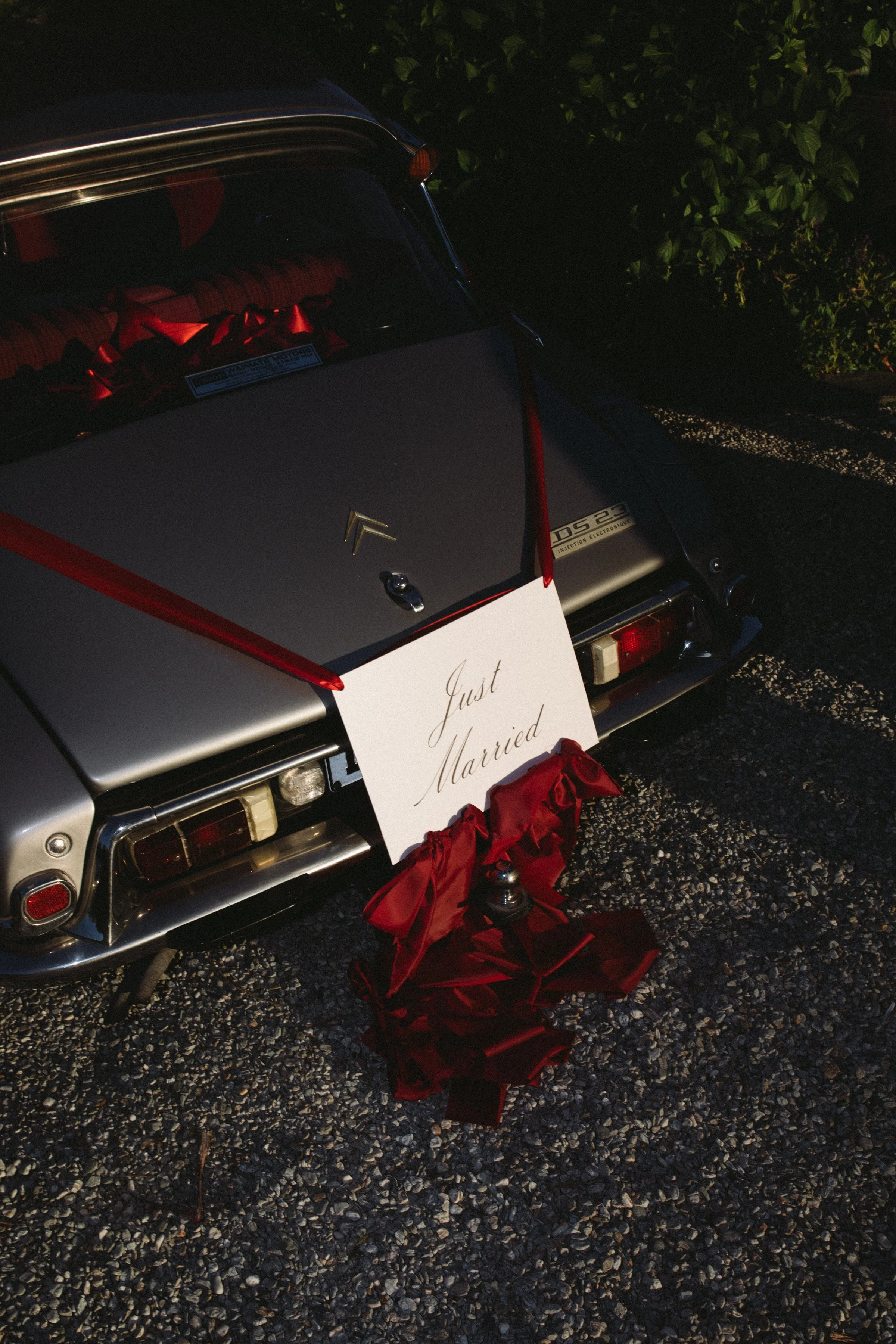 A classic silver car decorated for a wedding, with a sign that says 'Just Married', red ribbons, and red satin fabric on the ground behind it. The rear window has red ribbons, and there is greenery to the right.