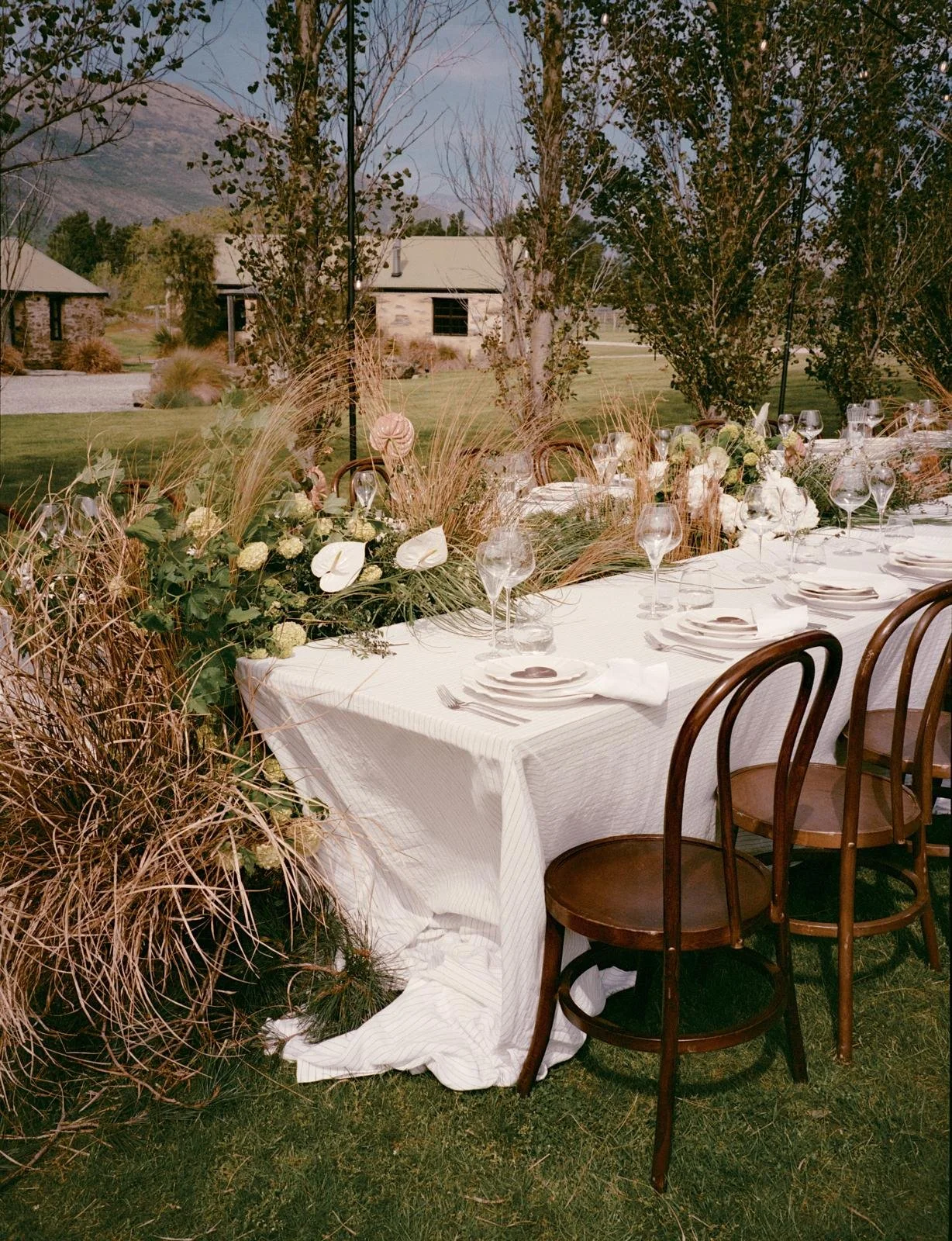 Elegant outdoor dining table decorated with floral centerpiece, white tablecloth, glassware, and place settings on a grassy lawn with trees and buildings in the background.