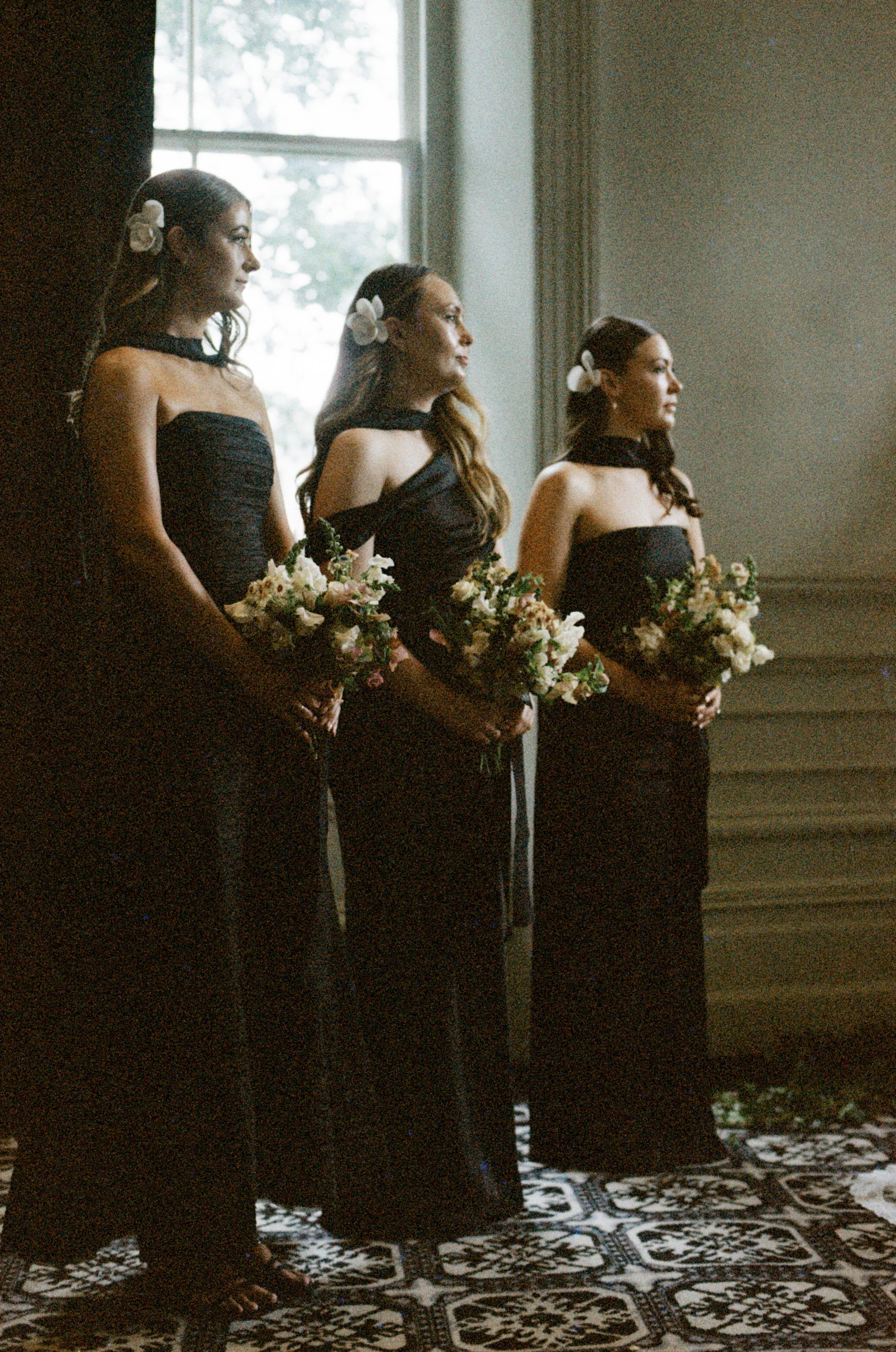 Three women are standing in a row indoors, dressed in black dresses with floral accessories in their hair, holding bouquets, with a patterned carpet on the floor and a window in the background.