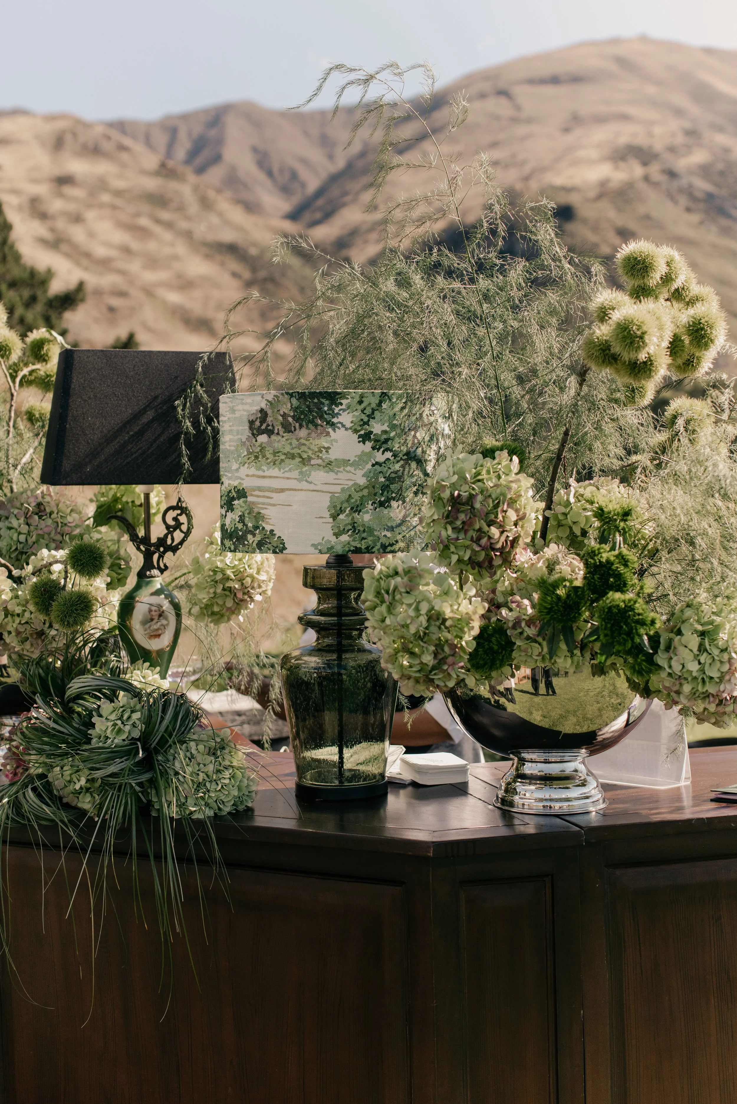 Various floral arrangements with white and green flowers and greenery, a decorative table lamp with a scenic landscape shade, placed on a dark wooden table outdoors with mountainous landscape in the background.