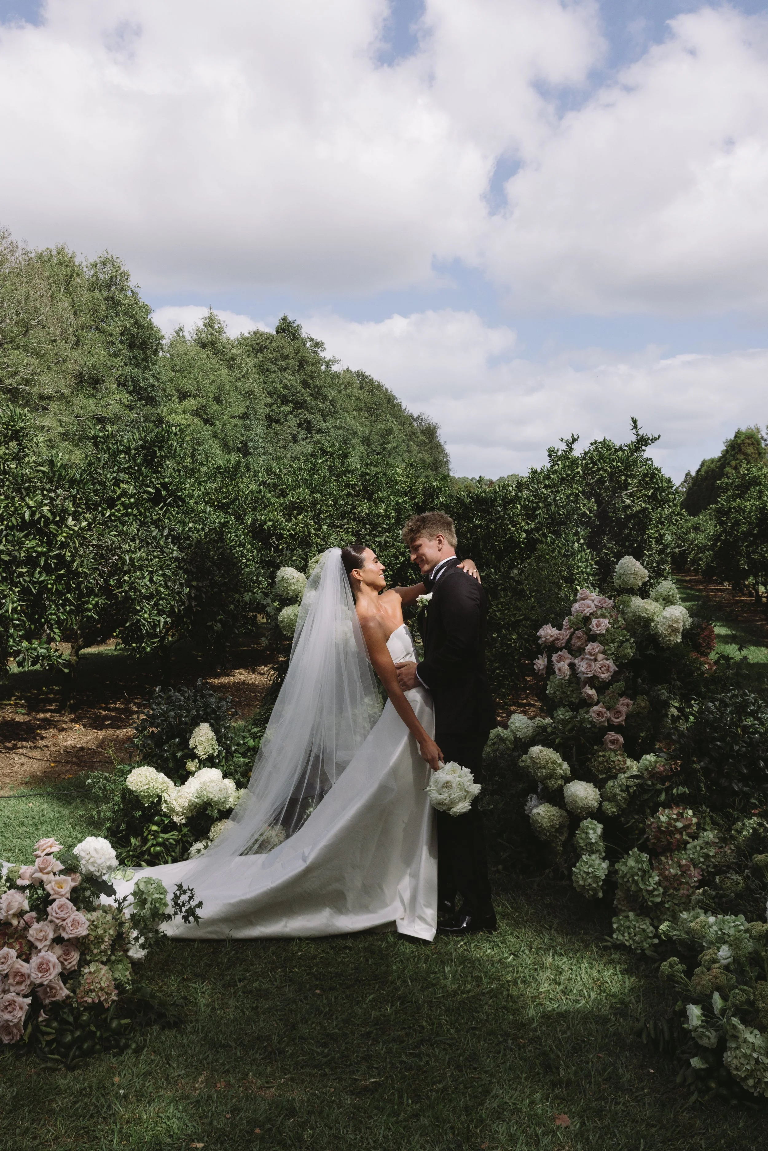 A bride and groom are sharing a moment outdoors during their wedding ceremony. The bride is wearing a white wedding dress with a veil, and the groom is in a black tuxedo. They are surrounded by lush green bushes and floral arrangements of white and pink roses. The sky above is partly cloudy.