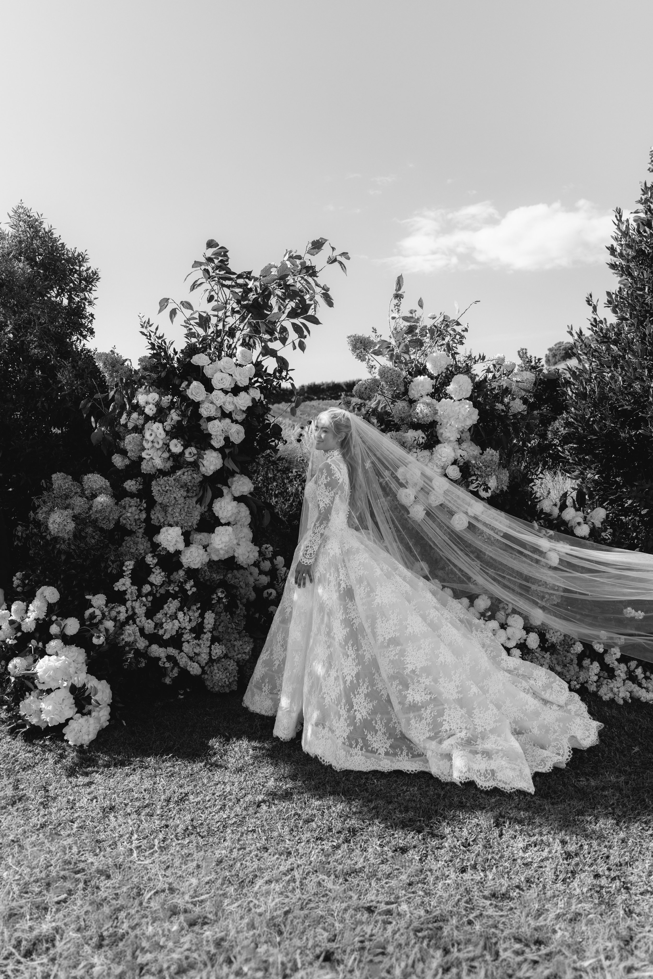 Black and white photo of a bride in a lace wedding dress with a long veil, standing outdoors among large flower arrangements and greenery, under a partly cloudy sky.