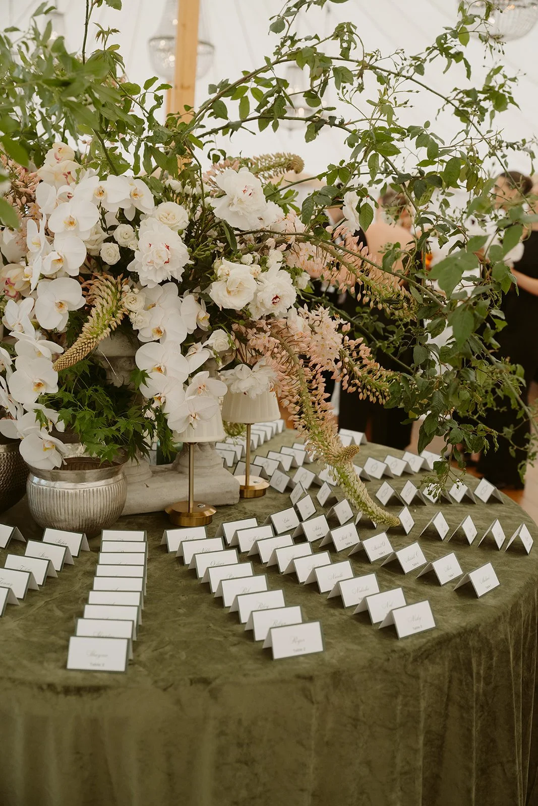 Table decorated with large floral arrangement of white orchids, peonies, and other flowers, surrounded by table name cards, at an event or wedding reception.