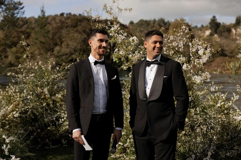 Two men in tuxedos with bow ties standing outdoors in front of flowering bushes and trees during daytime, possibly at a wedding or formal event.