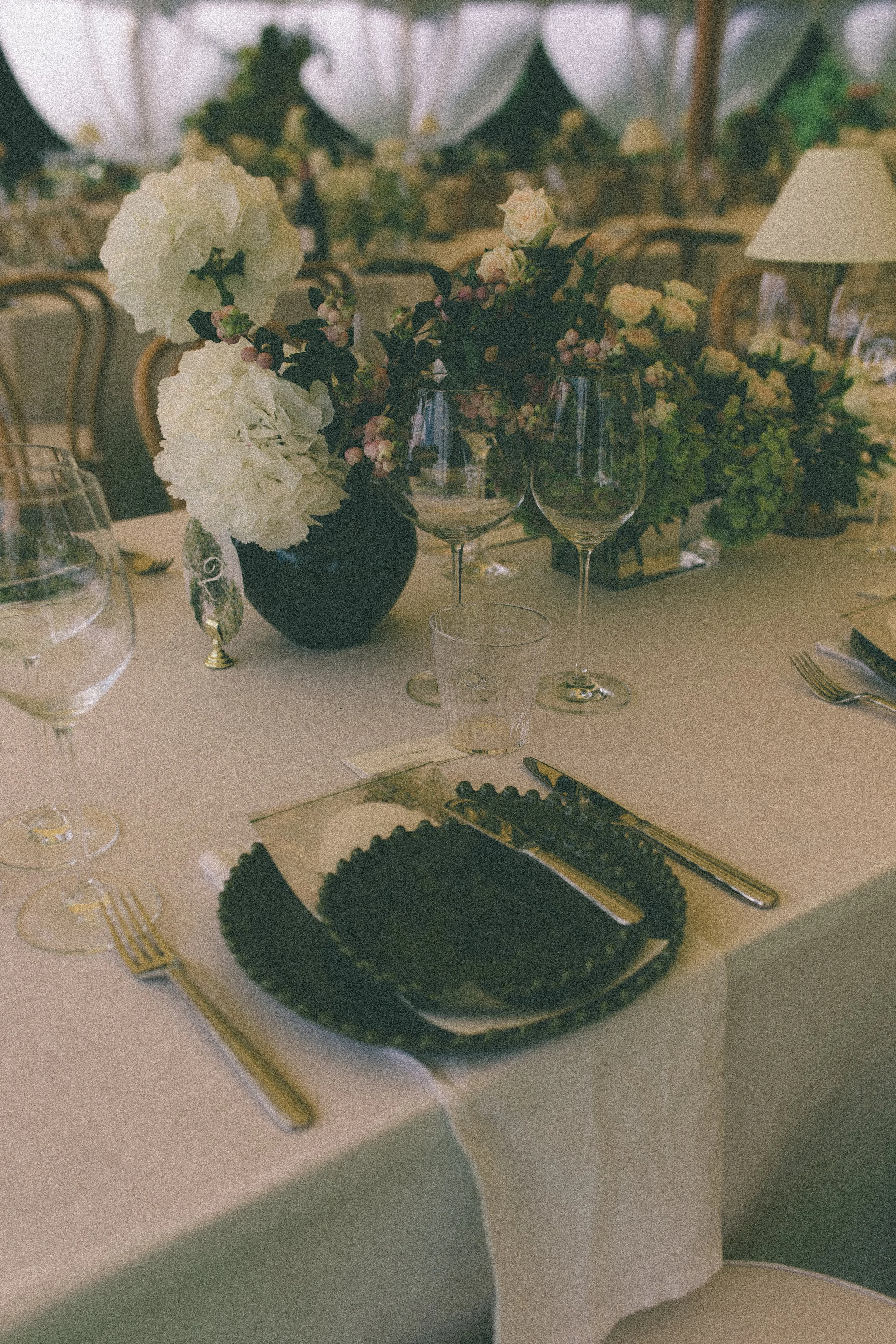 A table setting with a black dish with a gold-rimmed edge, gold utensils, a wine glass, a water glass, and a decorative floral centerpiece with white and pink flowers in a black vase. The background shows a dining area with chairs and additional floral arrangements.