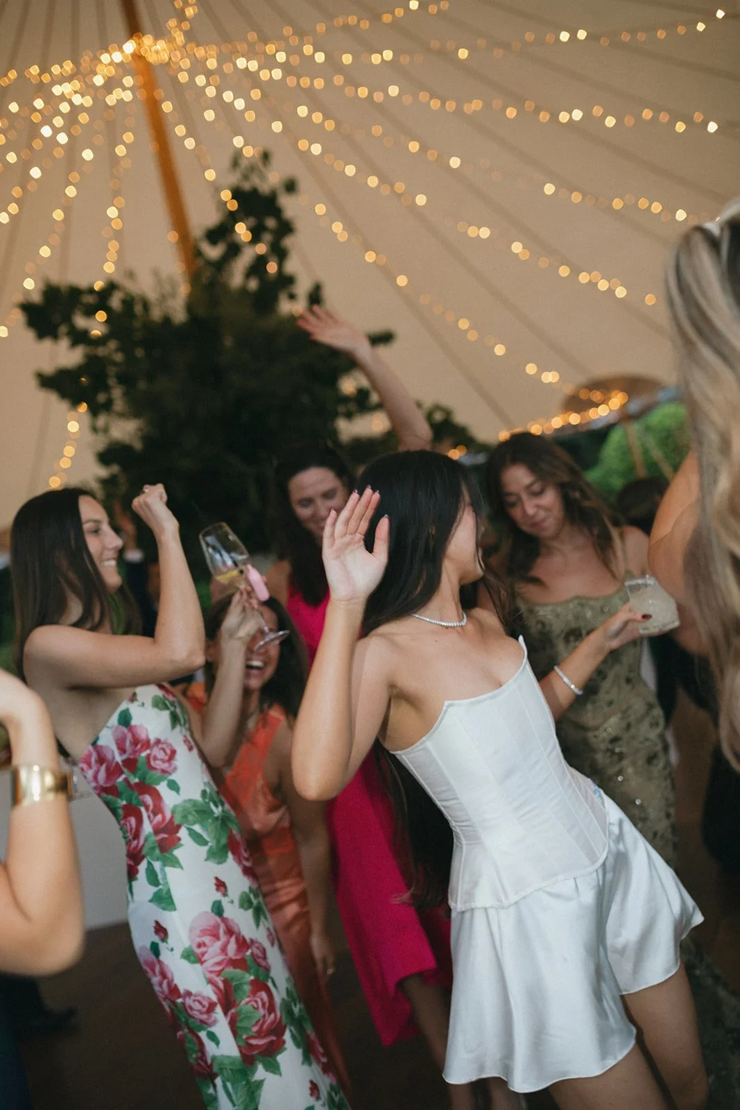 Group of women dancing and celebrating at a party under string lights, with some holding drinks.