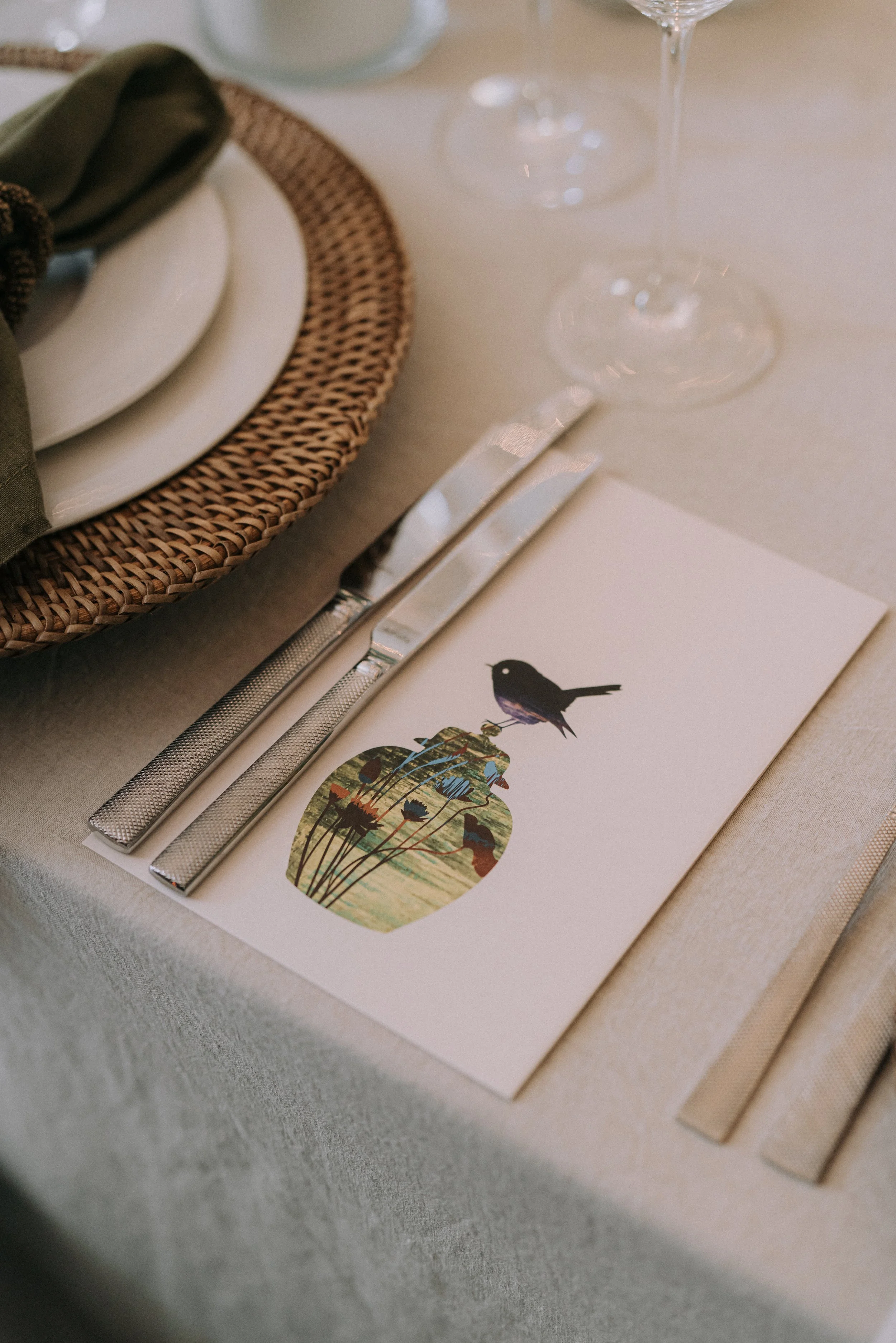 Close-up of a table setting with a woven placemat, white plate, silverware, two empty wine glasses, and a menu with a colorful illustration of a bird and plants.