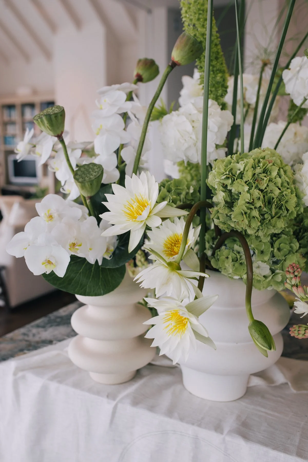 Arrangement of white orchids, green hydrangeas, and white water lilies in white ceramic vases on a table.