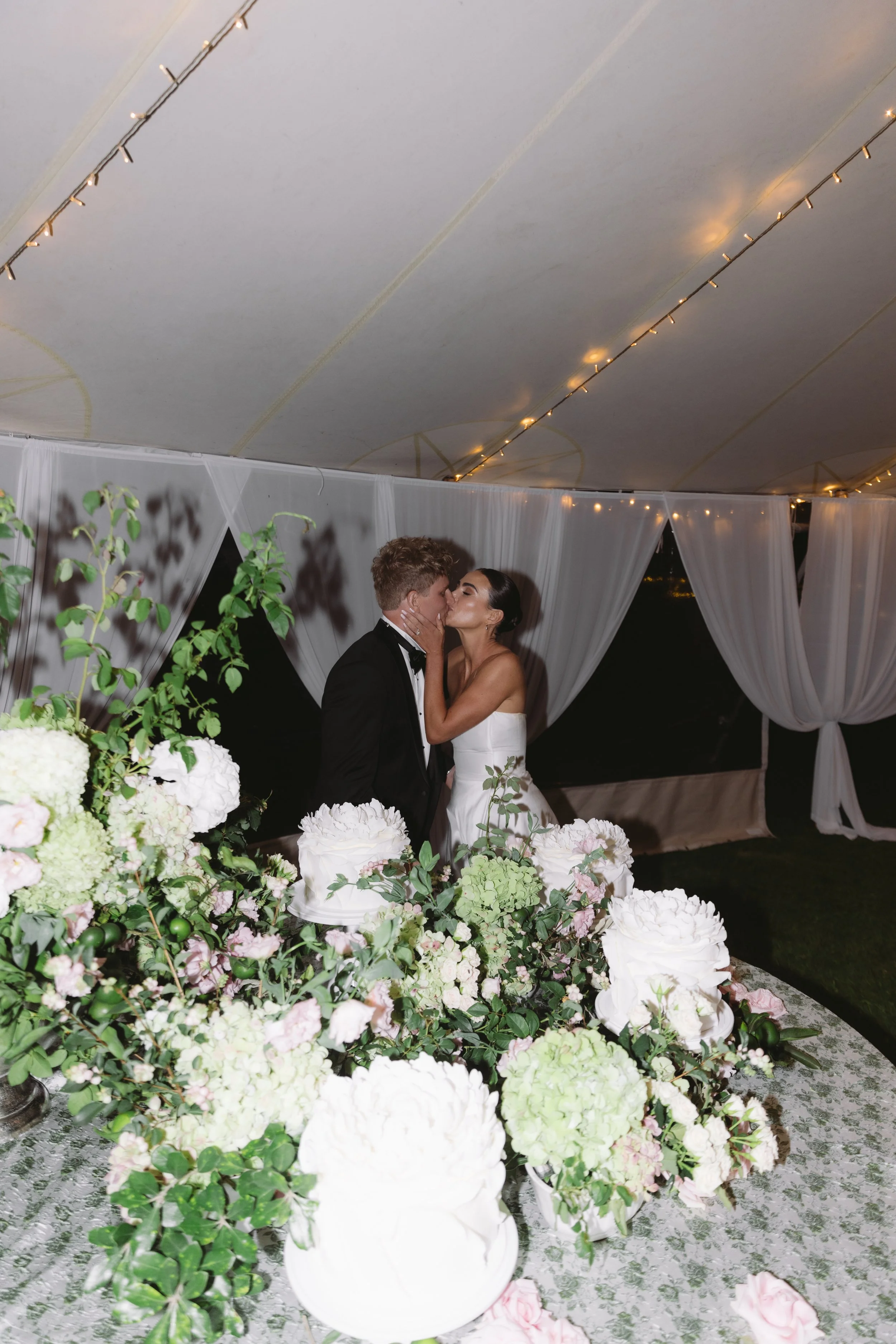 A bride and groom kiss at their wedding reception under a draped canopy with string lights. The scene is decorated with white and pastel-colored flowers.