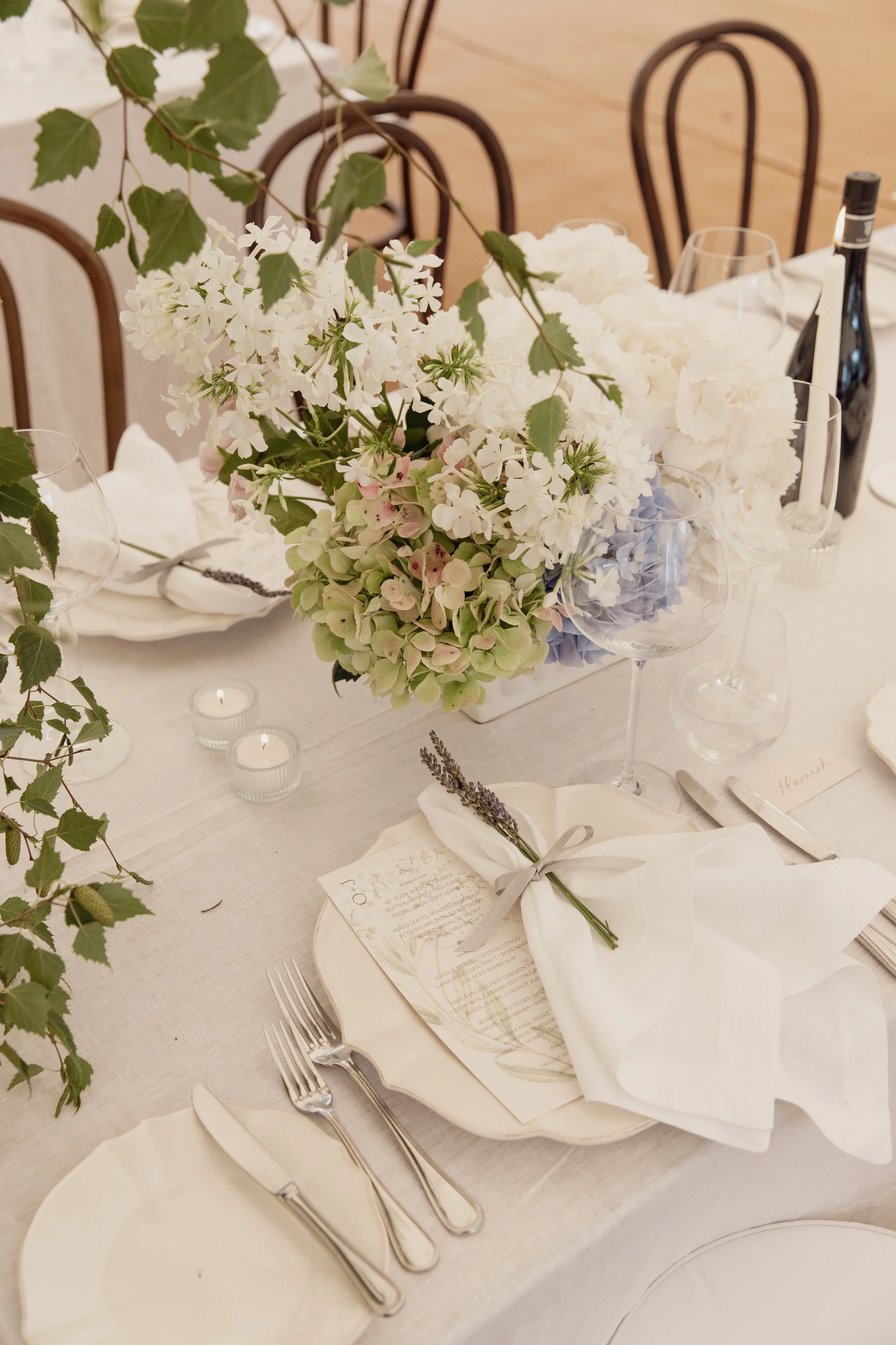 A table set for a formal event with a floral centerpiece, white napkins, silverware, candles, a wine bottle, and glassware. The centerpiece includes white and light pastel flowers and green foliage. The table has a white tablecloth and each place setting features a menu card and a small sprig of lavender.