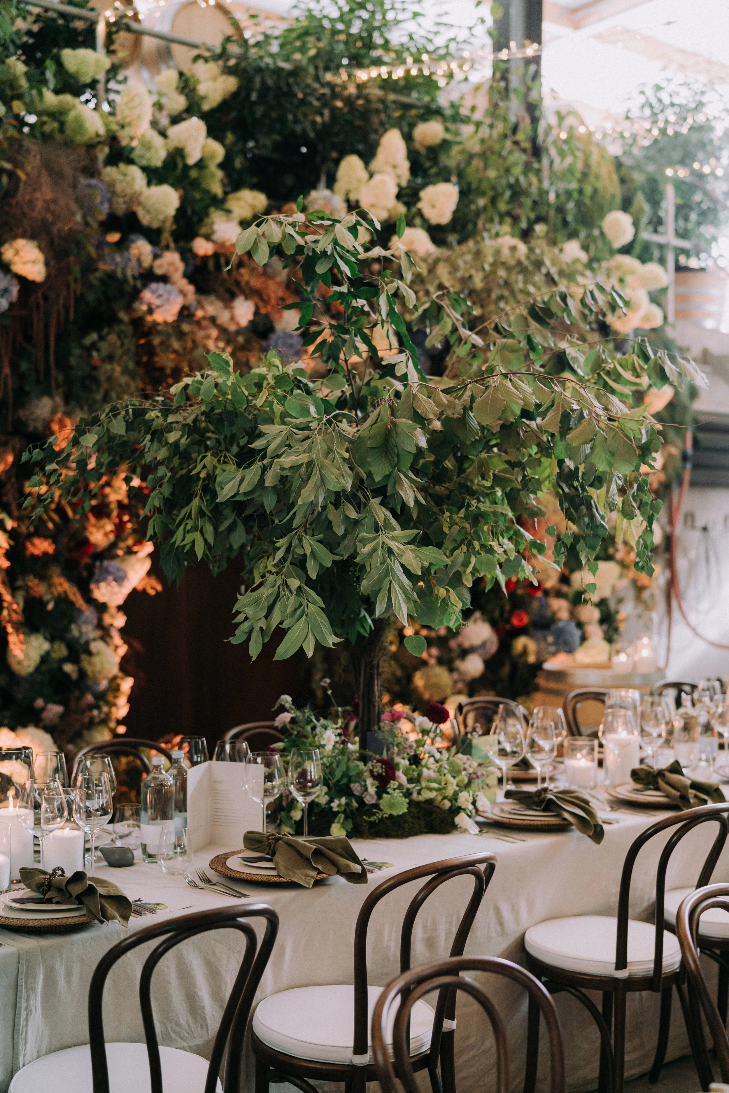 A decorated dining table at an event with a tall centerpiece of green leaves, surrounded by floral arrangements, glasses, and place settings, with a floral and string light backdrop.