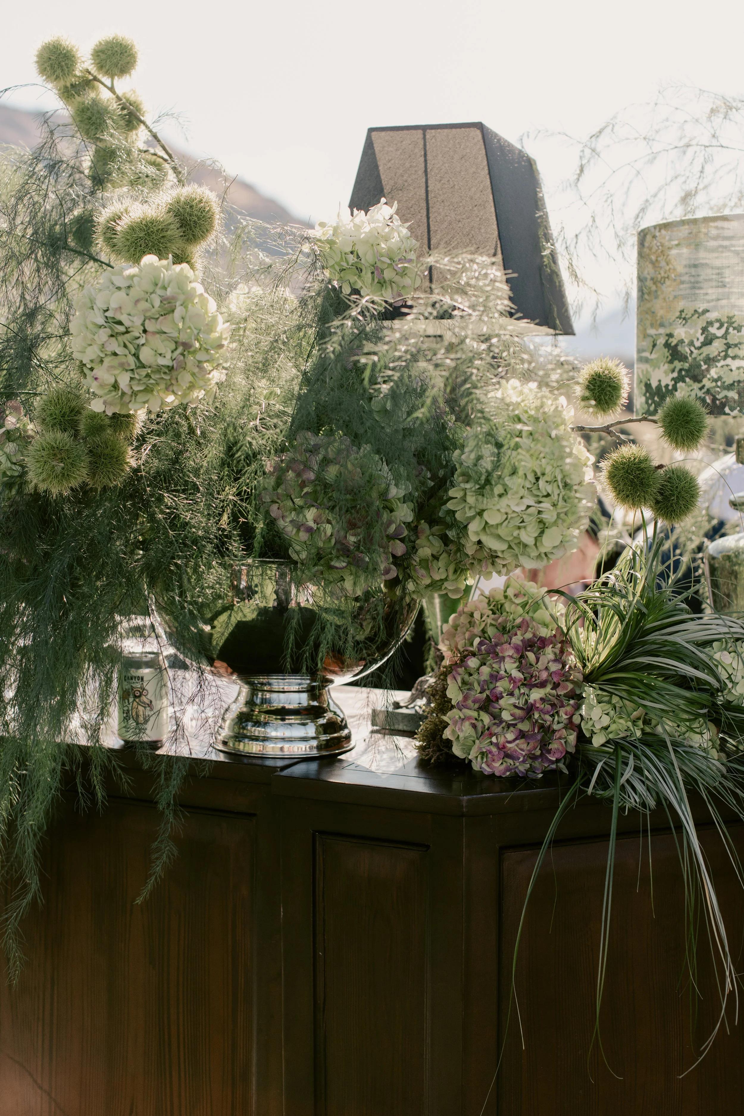 A floral arrangement featuring white and purple hydrangeas, green thistle, and various greenery in a silver vase on a dark wooden cabinet.