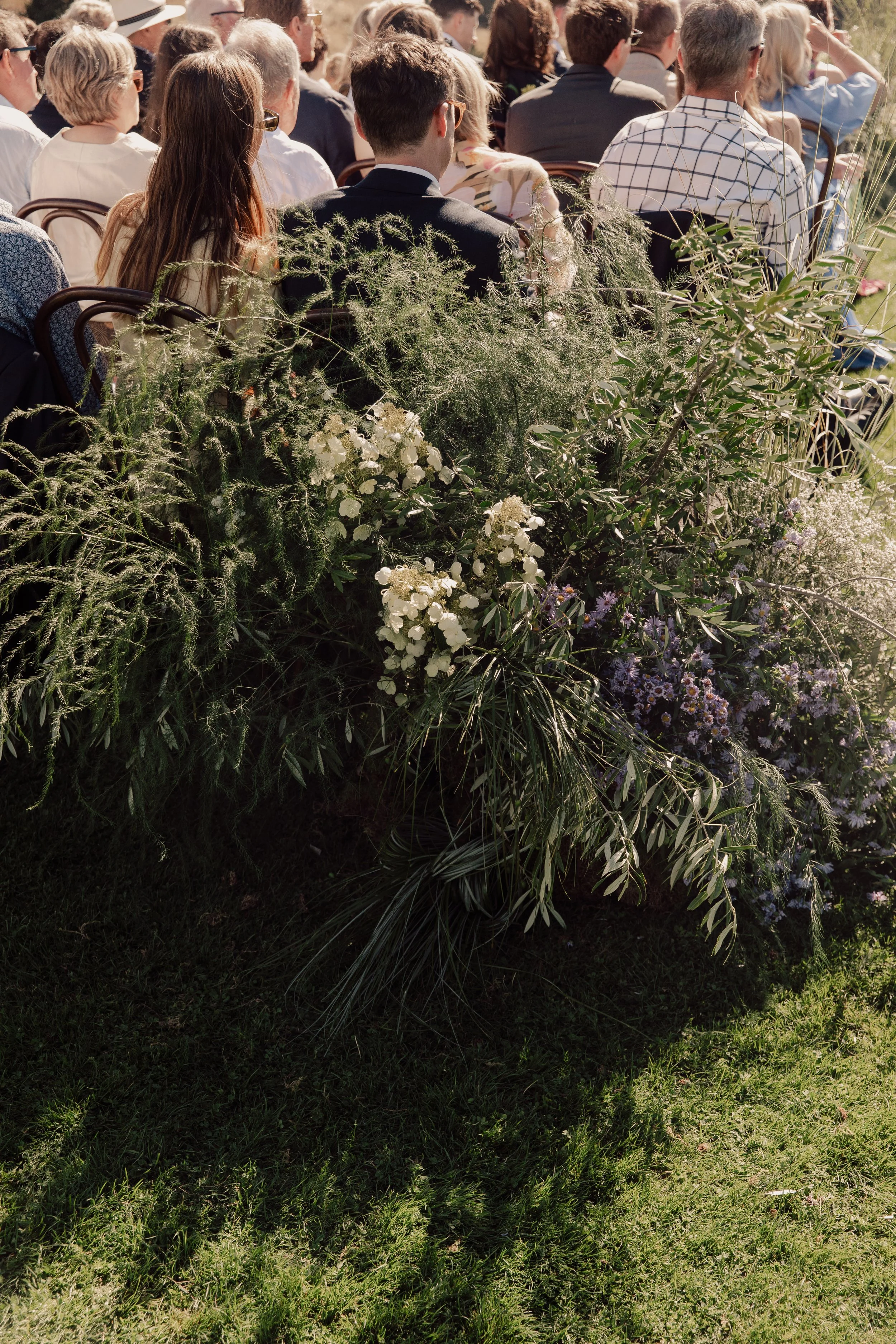 A crowd of people sitting outdoors at a wedding ceremony on chairs, facing away from the camera, during a sunny event. There are green plants and flowers in the foreground.