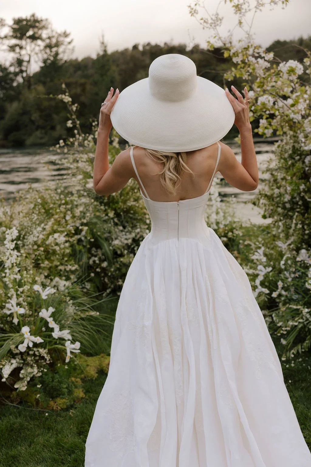 A woman in a white dress and large sunhat standing outdoors amid flowering bushes and greenery, facing away from the camera.