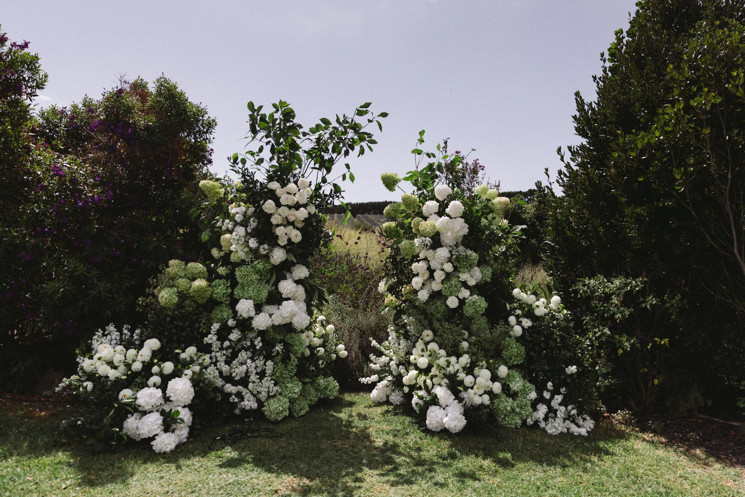 An outdoor floral display with white and green hydrangeas, and other plants, set against a background of trees and a pale sky.