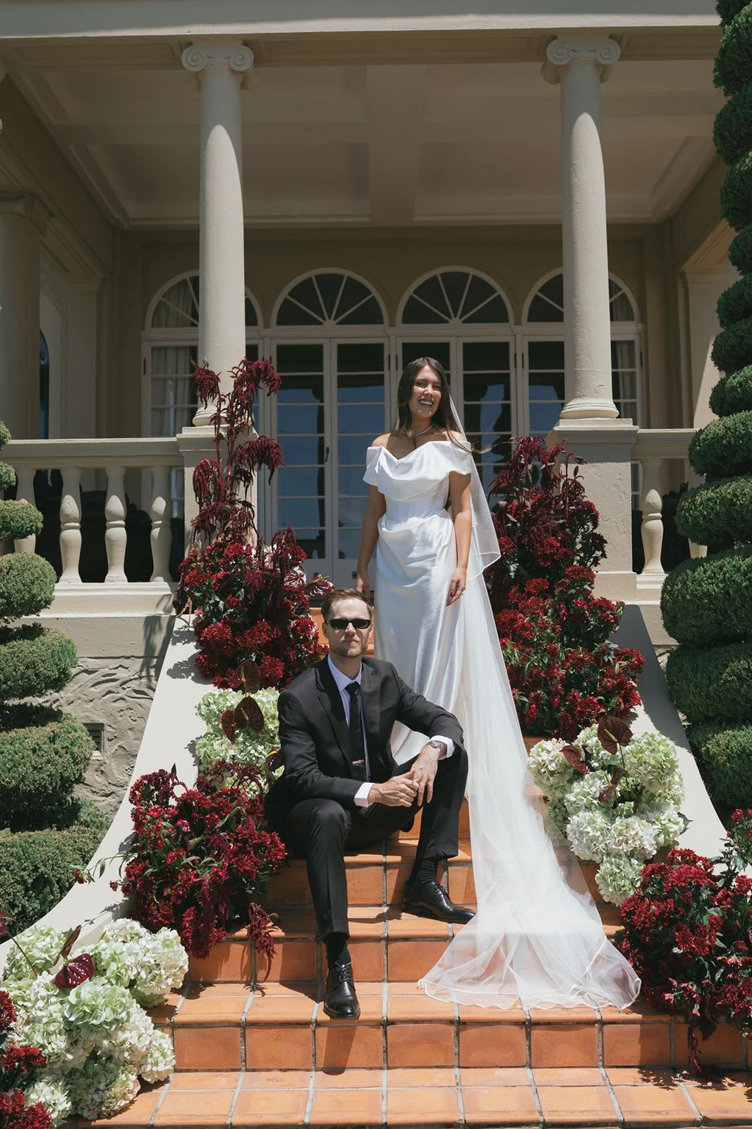 Bride in a white wedding dress and groom in a black suit with sunglasses on the front steps of a large house, surrounded by red and white flowers and greenery.