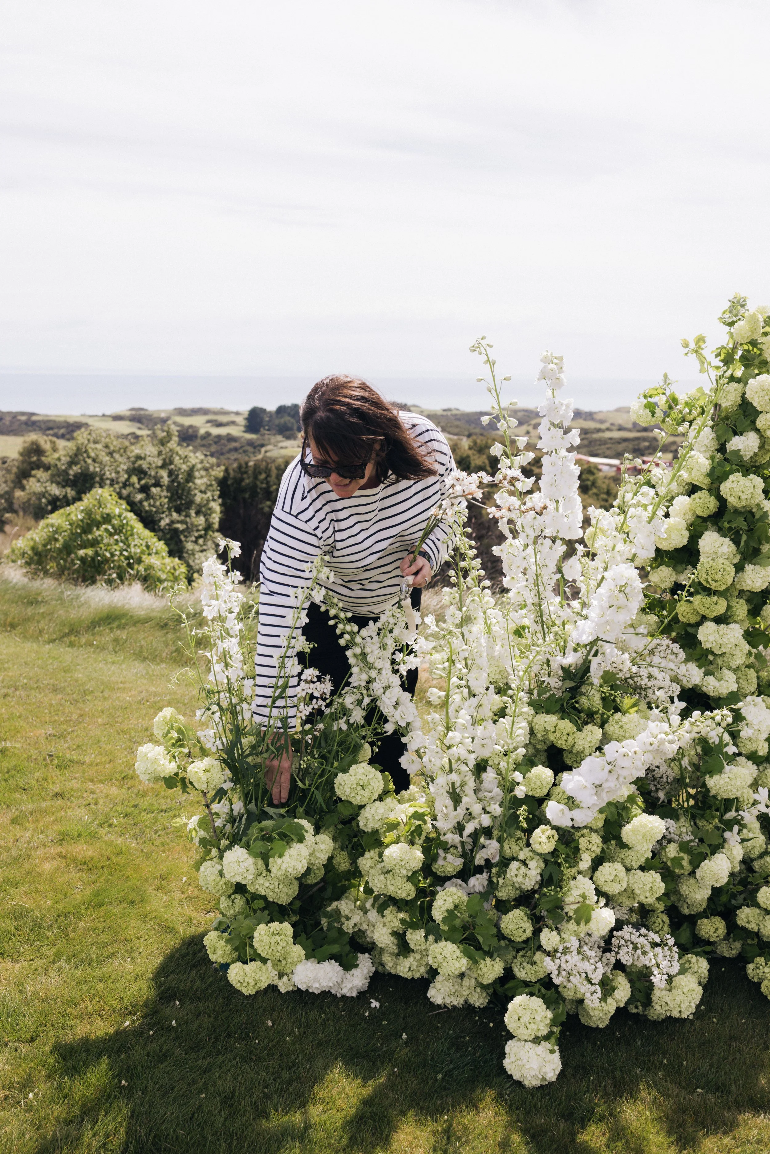 A woman in sunglasses and a striped shirt bends down to tend to white hydrangeas and delphiniums in a garden, with rolling hills and a view of the ocean in the background under a partly cloudy sky.