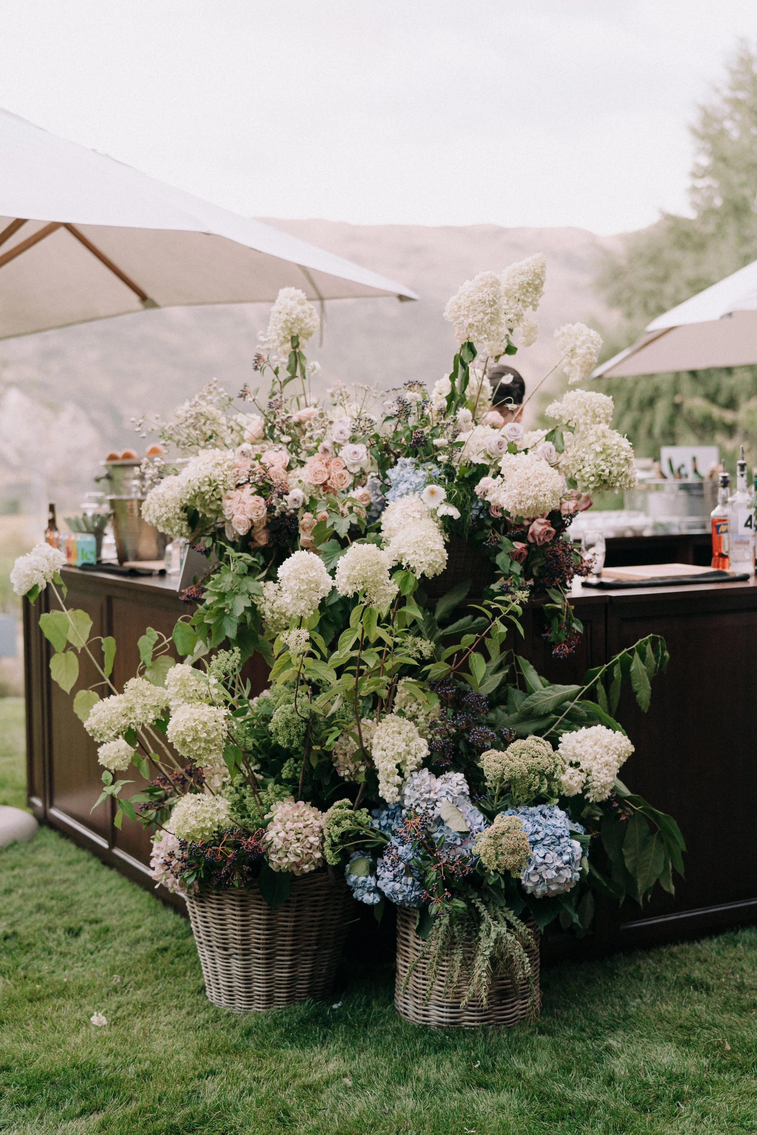 Large floral arrangement with white, pink, and blue hydrangeas, roses, and greenery in wicker baskets, set outdoors with umbrellas and mountains in the background.
