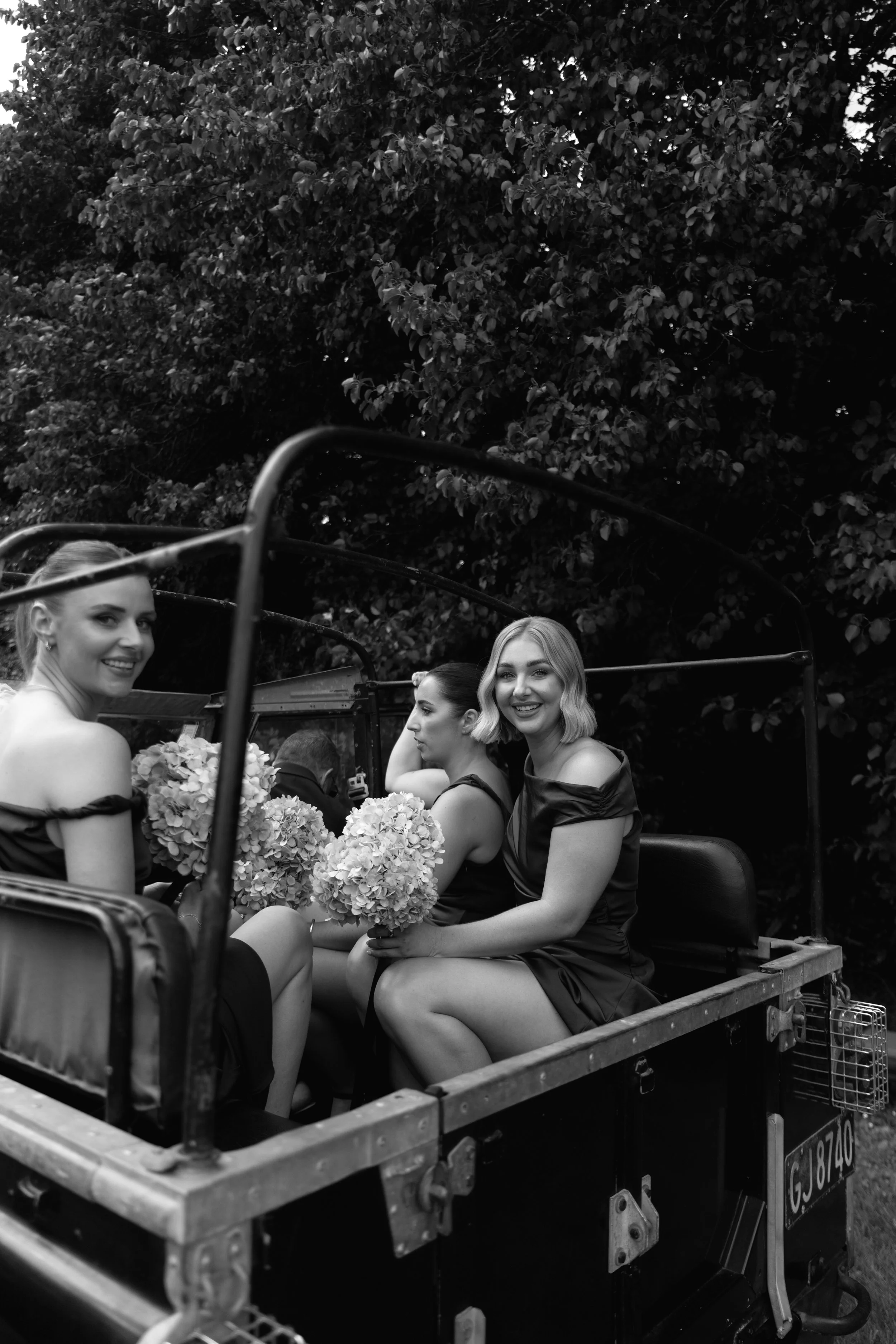 Three women sitting in the back of a vintage truck, holding bouquets of flowers, with trees in the background. The women are smiling and dressed in elegant dark dresses, suggesting they are at a celebration or event.