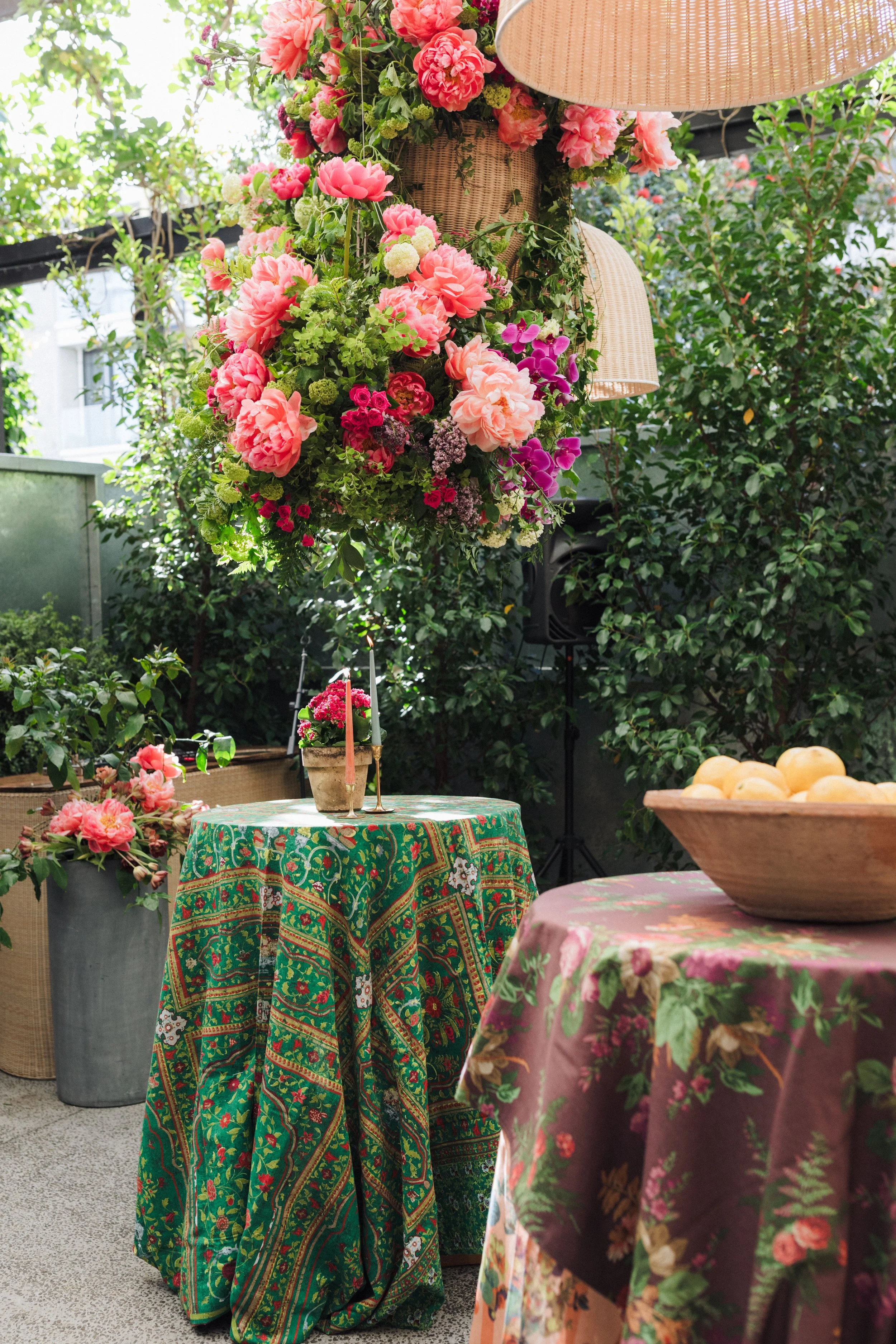 A garden setting with tables covered in vibrant, patterned tablecloths, and surrounded by lush greenery and blooming flowers. A large hanging floral arrangement in a woven basket is prominent, with pink, green, and purple blossoms. There are also candles and potted plants on the tables.