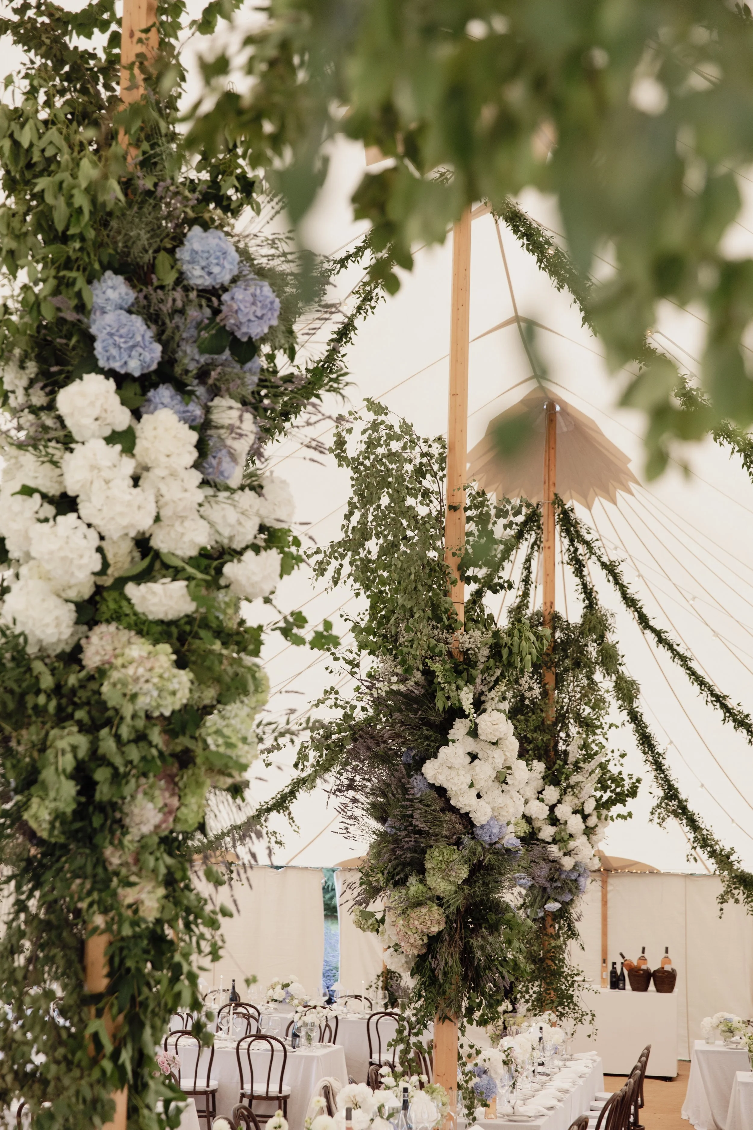 A decorated event tent with white drapery, green foliage, and a floral arrangement featuring white and blue hydrangeas, set for a formal celebration.