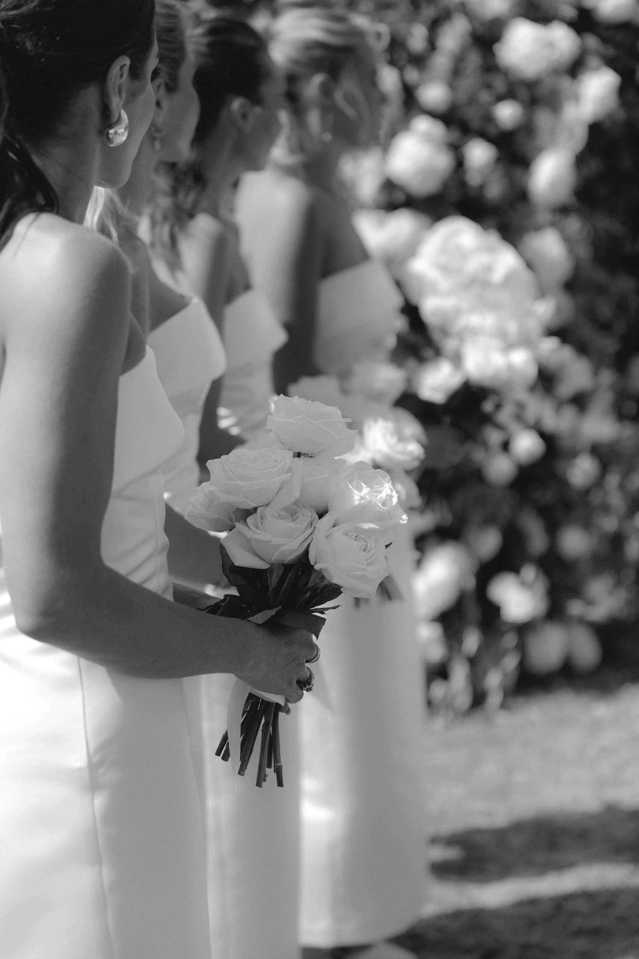 Four women in white dresses holding bouquets of white roses, standing in a line outdoors.
