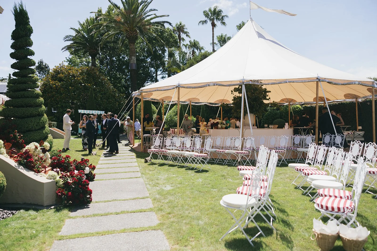 People gathered outdoors at a wedding reception with a large white tent, decorative chairs with striped cushions, and lush greenery including tall trees and blooming bushes.